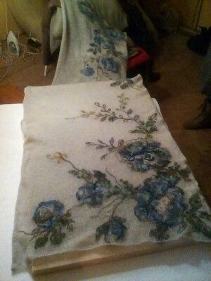 Dwa kawałki jasnego jedwabiu z ręcznie malowanymi motywami kwiatowymi w odcieniach niebieskiego i zieleni, leżące na stole w słabo oświetlonym pomieszczeniu, z żelazkiem widocznym w tle.