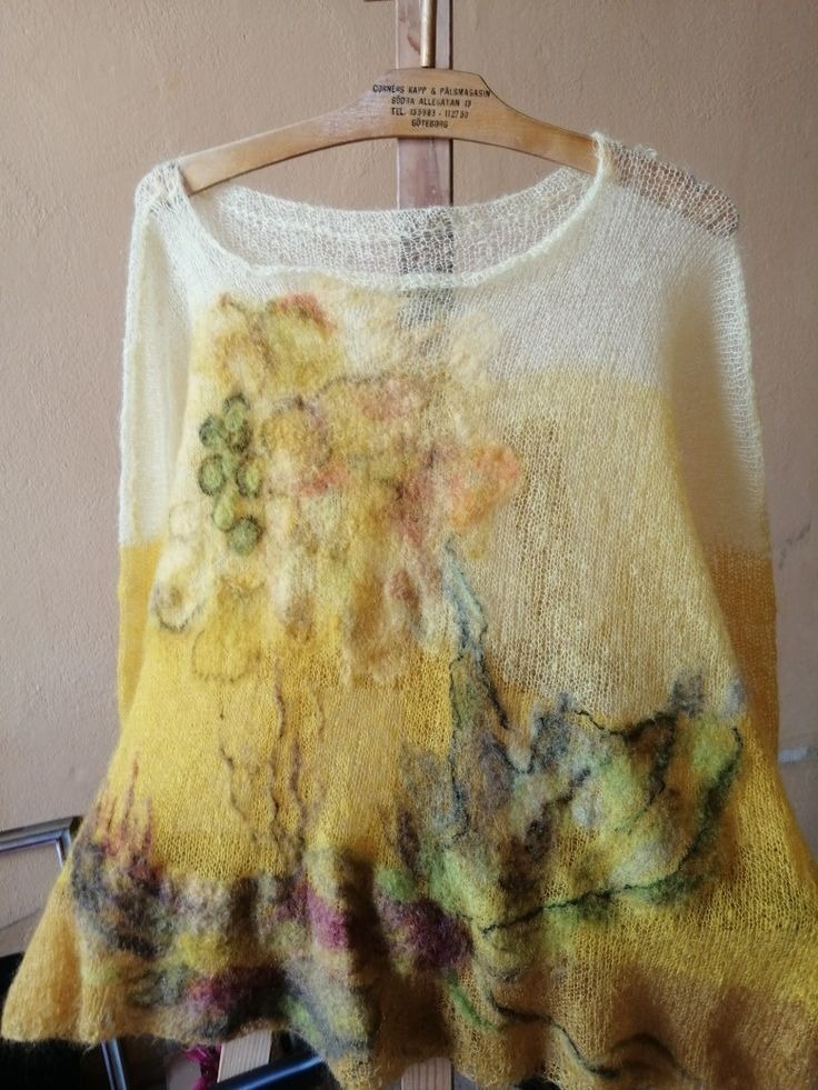 Żółty, ażurowy sweter z ręcznie filcowanymi aplikacjami kwiatowymi, zawieszony na drewnianym wieszaku z napisem 'Corners Kapp & Pälsmagasin'.