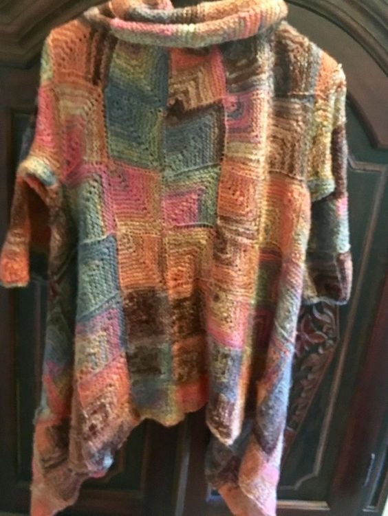 Ręcznie robiona tunika z grubym golfem, wykonana z kolorowej włóczki w kwadraty, widoczna na tle ciemnych drzwi z ornamentami.