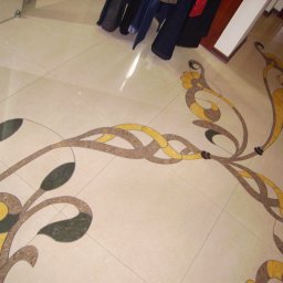 Olek Jaworski - Beżowa podłoga z płytek ceramicznych z dekoracyjnym, roślinnym wzorem w odcieniach brązu, żółci i zieleni, widoczne ubrania i fragment buta.