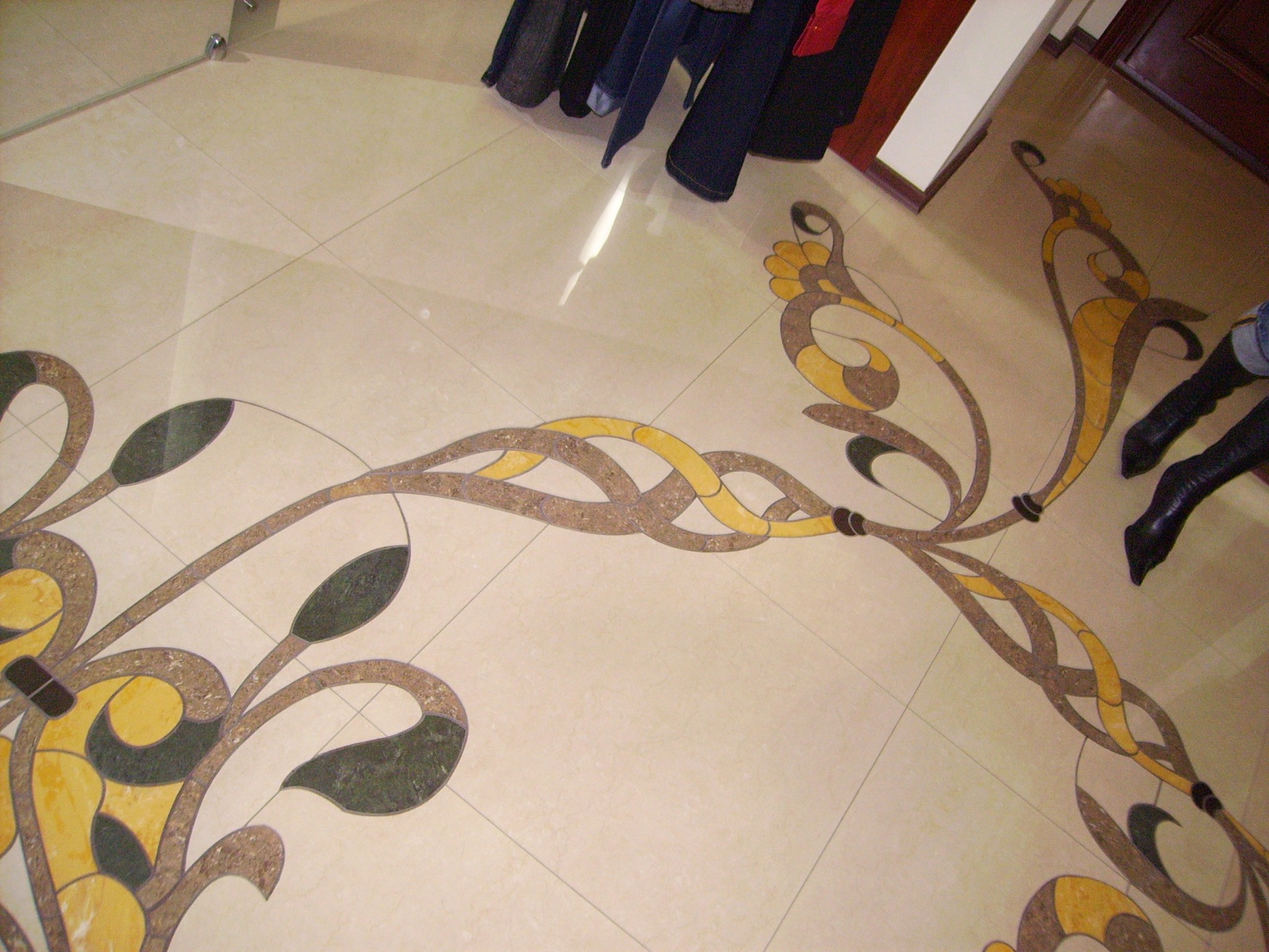 Beżowa podłoga z płytek ceramicznych z dekoracyjnym, roślinnym wzorem w odcieniach brązu, żółci i zieleni, widoczne ubrania i fragment buta.