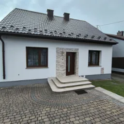 Elewacja domu jednorodzinnego po remoncie: białe ściany, brązowy dach z dachówki, kamienna okładzina wokół drzwi, kostka brukowa na podjeździe.
