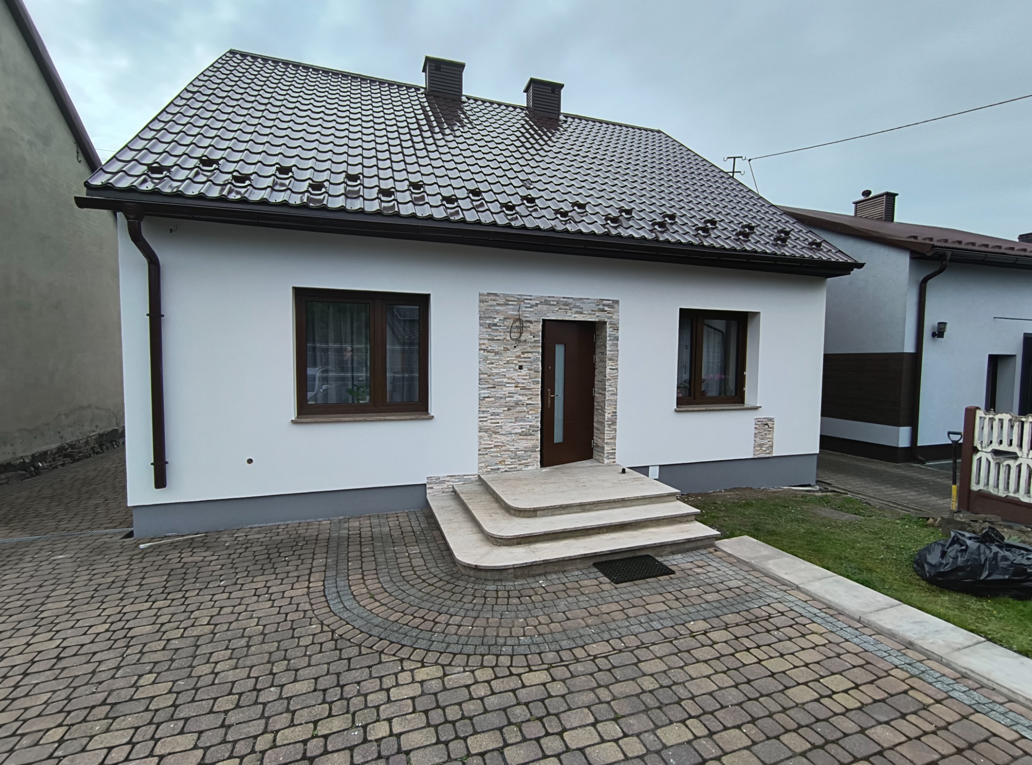 Elewacja domu jednorodzinnego po remoncie: białe ściany, brązowy dach z dachówki, kamienna okładzina wokół drzwi, kostka brukowa na podjeździe.