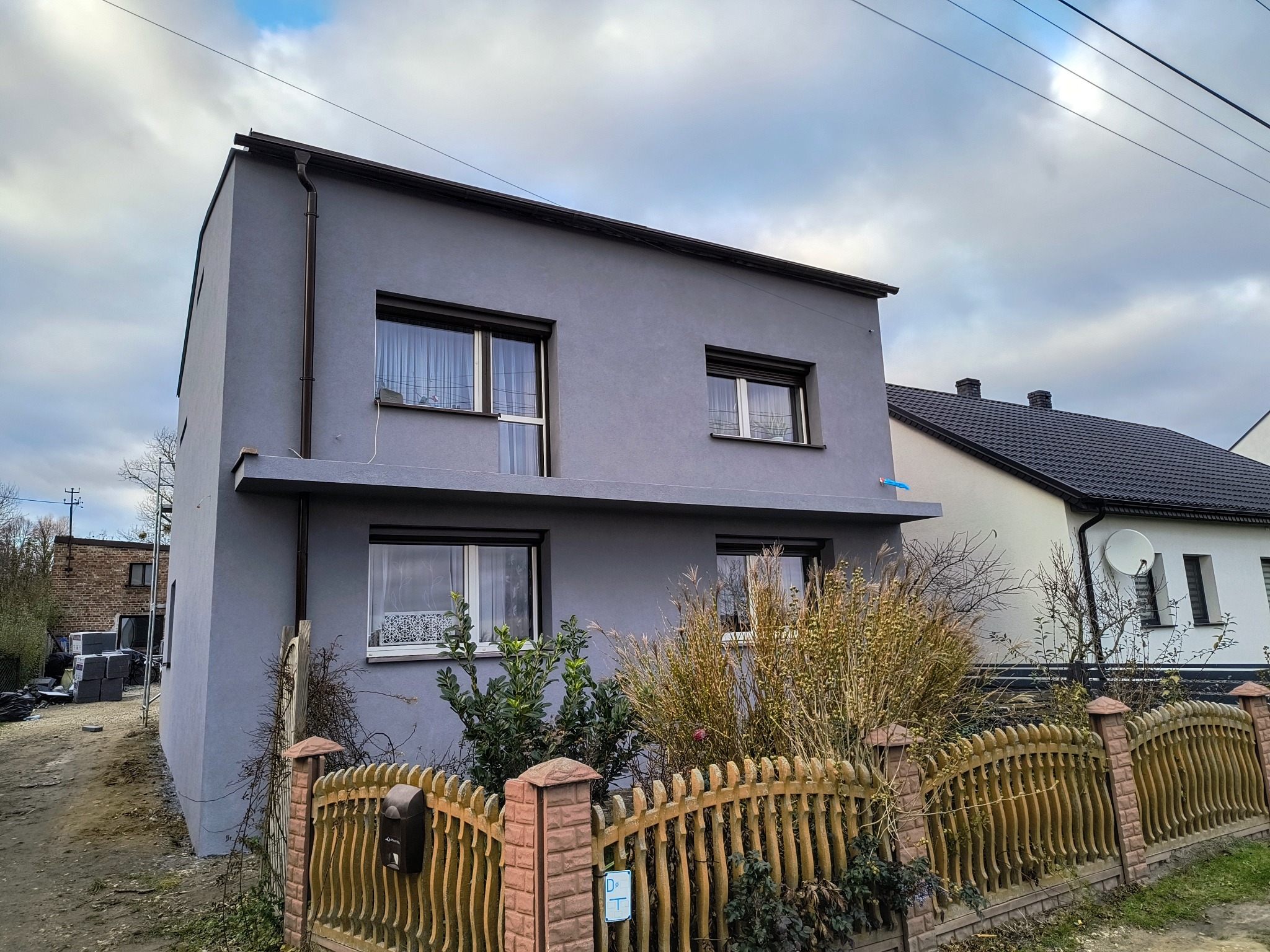 Szary, dwukondygnacyjny dom jednorodzinny z brązowymi oknami i dachem, ogrodzony drewnianym płotem. Widoczne elementy elewacji i zagospodarowania terenu.