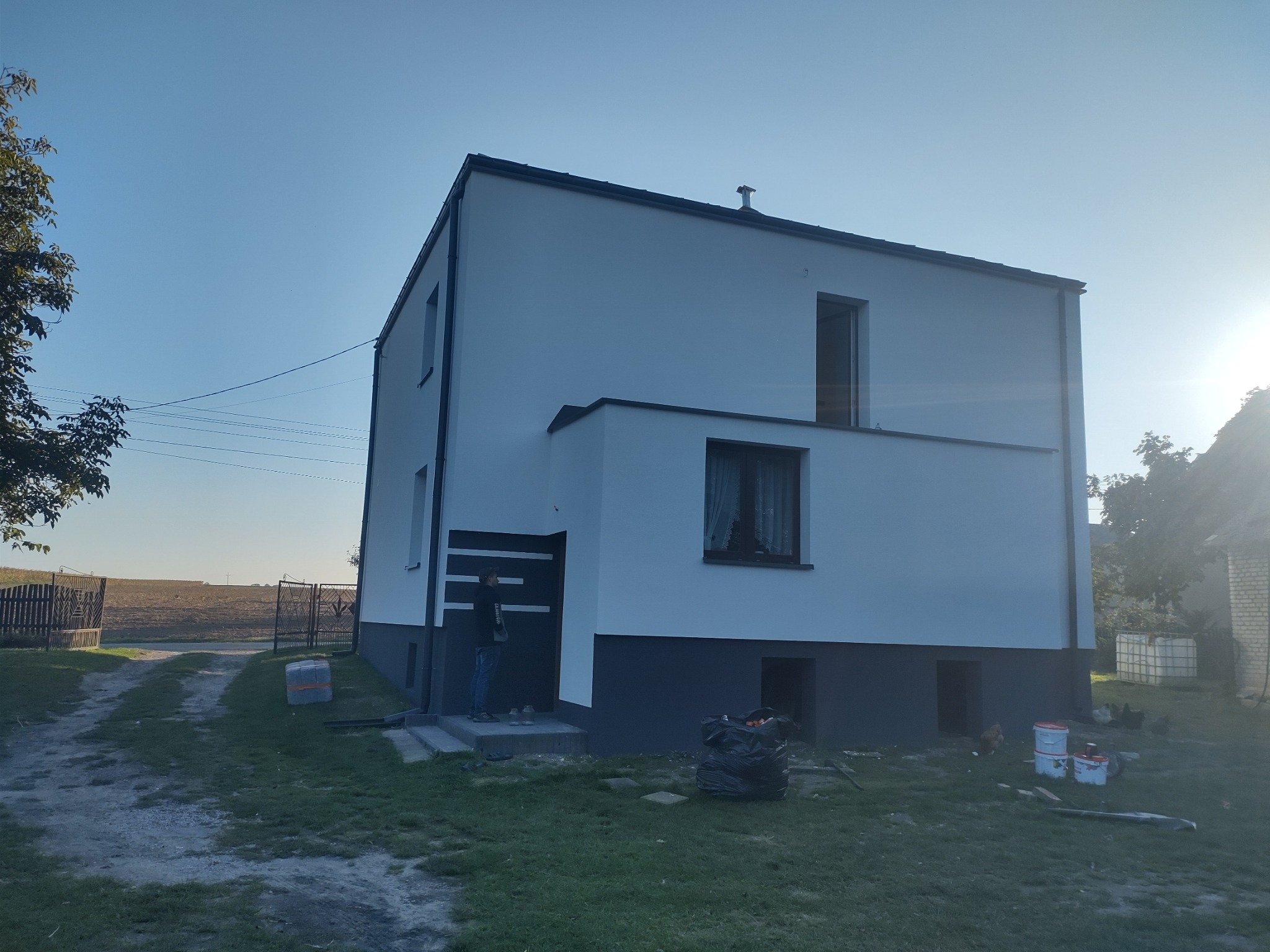 Dwukondygnacyjny dom z białą elewacją i antracytowymi detalami. Widoczna osoba przed wejściem i puszki po farbie na trawniku. Słoneczny dzień.