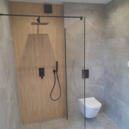 Mid-Ar Wykończenia Wnętrz - Nowoczesna łazienka z kabiną prysznicową typu walk-in, wykończona płytkami imitującymi marmur i drewno, z czarną armaturą i podtynkową miską WC.