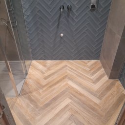 Mid-Ar Wykończenia Wnętrz - Wykończona łazienka z drewnopodobną podłogą ułożoną w jodełkę i szarymi płytkami na ścianie, również w jodełkę, widoczna szklana kabina prysznicowa i fragment instalacji wodnej.