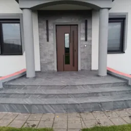 Wykończone schody wejściowe do domu z szarych płytek imitujących kamień, z ciemnobrązowymi drzwiami i szarą elewacją. Widoczne kolumny i numer domu.