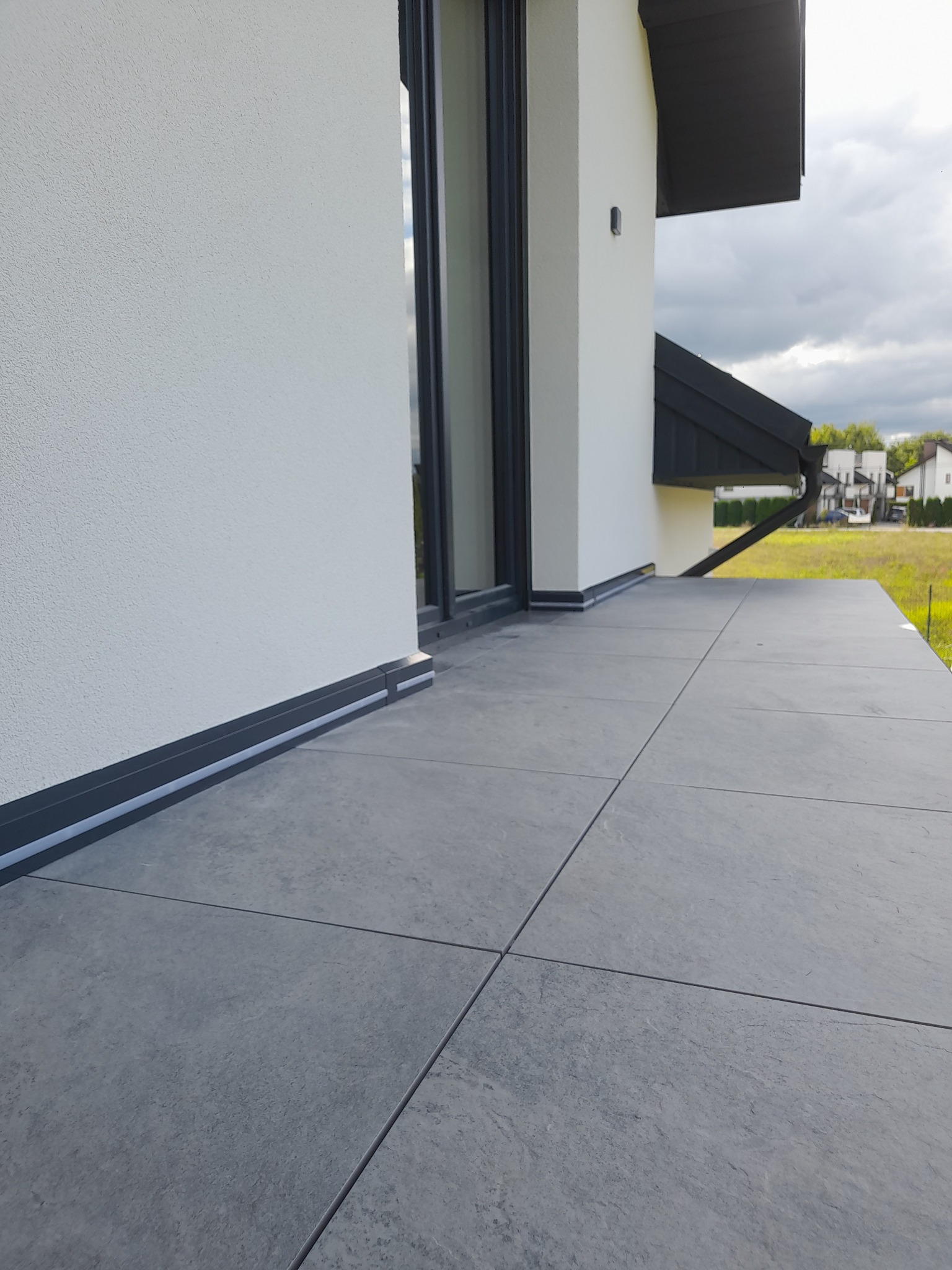 Szare płytki gresowe ułożone na tarasie domu jednorodzinnego, widok z zewnątrz, z widoczną elewacją budynku i fragmentem ogrodu w tle.