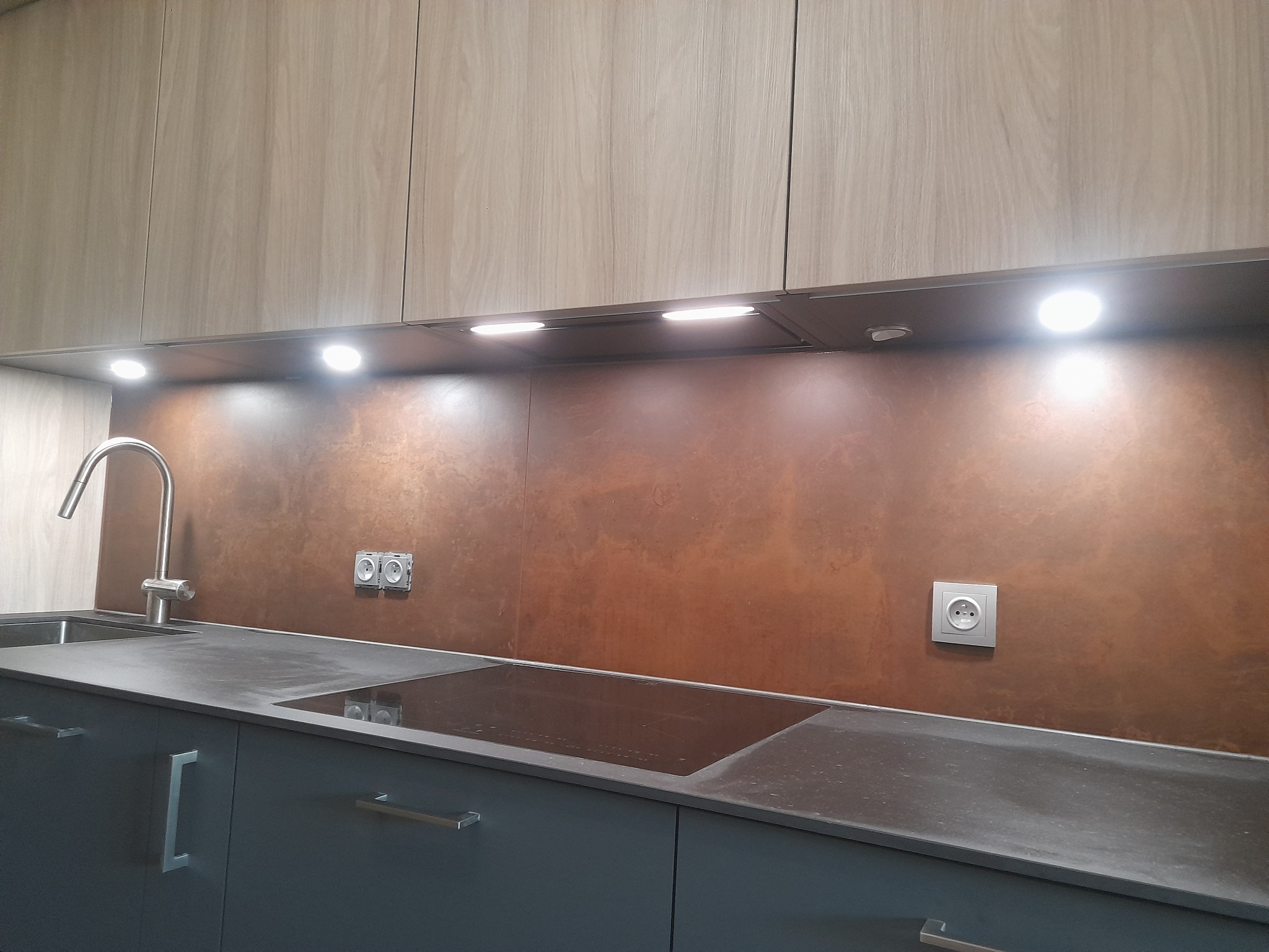 Nowoczesna kuchnia z szarymi szafkami, blatem w kolorze grafitu, miedzianą ścianą nad blatem i oświetleniem podszafkowym LED.