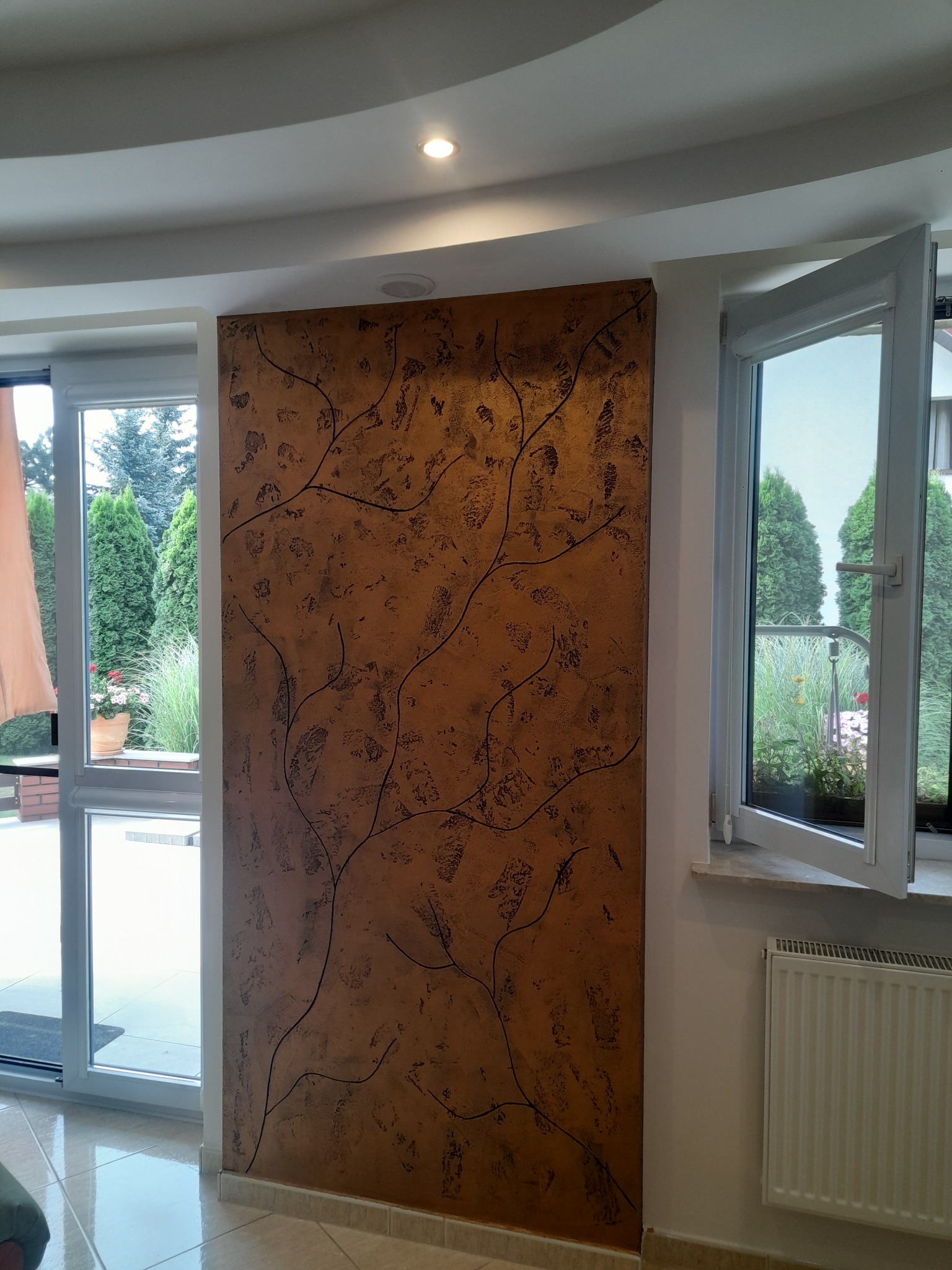 Ściana z dekoracyjną fakturą w odcieniach brązu, zdobiona motywem gałązek, sąsiadująca z oknem wychodzącym na ogród.