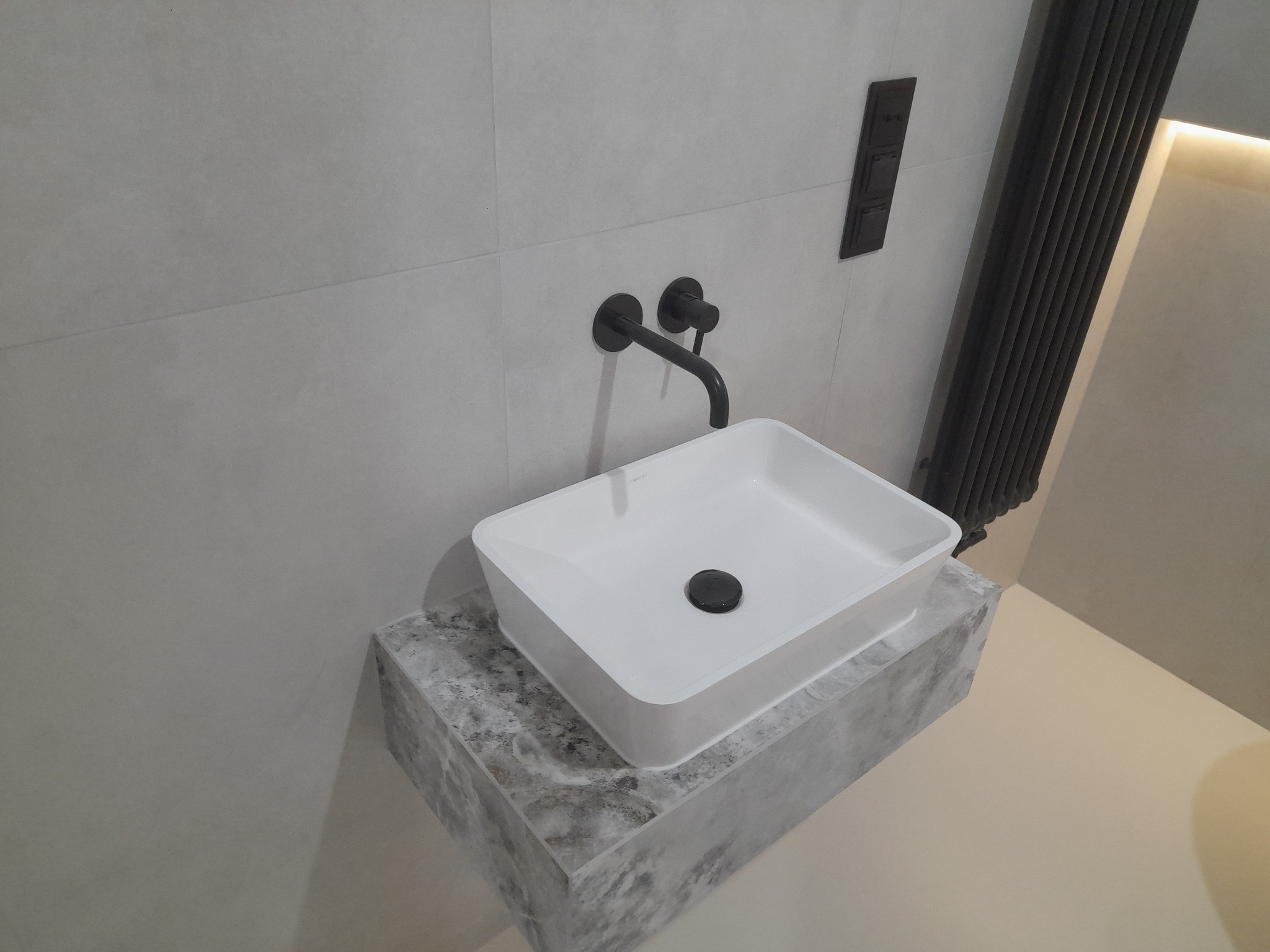 Nowoczesna umywalka nablatowa o kwadratowym kształcie, z czarną armaturą i odpływem, umieszczona na blacie imitującym marmur, na tle jasnych płytek i grzejnika żeberkowego w kolorze czarnym.