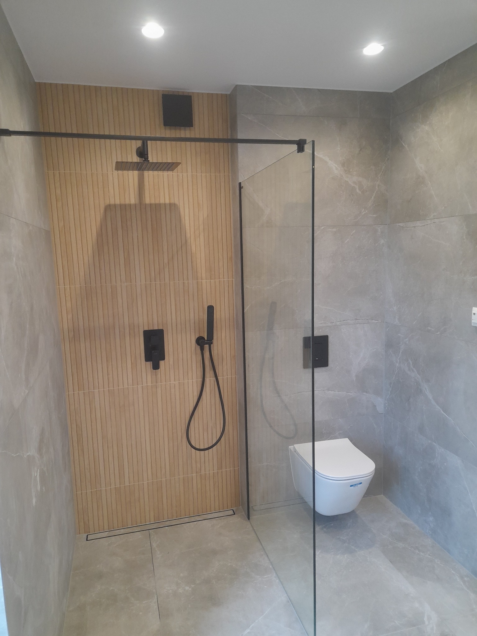 Nowoczesna łazienka z kabiną prysznicową typu walk-in, wykończona płytkami imitującymi marmur i drewno, z czarną armaturą i podtynkową miską WC.