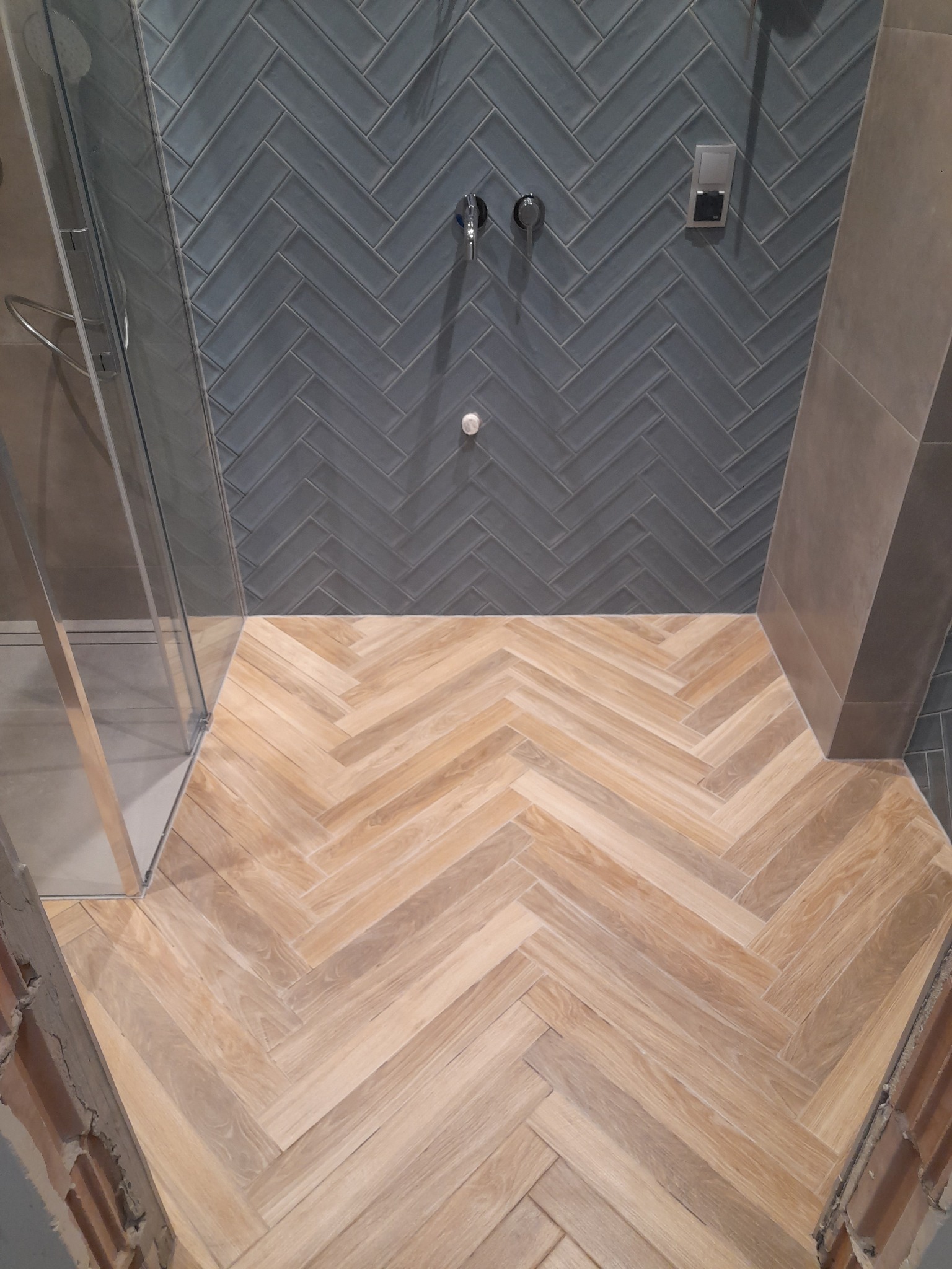 Wykończona łazienka z drewnopodobną podłogą ułożoną w jodełkę i szarymi płytkami na ścianie, również w jodełkę, widoczna szklana kabina prysznicowa i fragment instalacji wodnej.