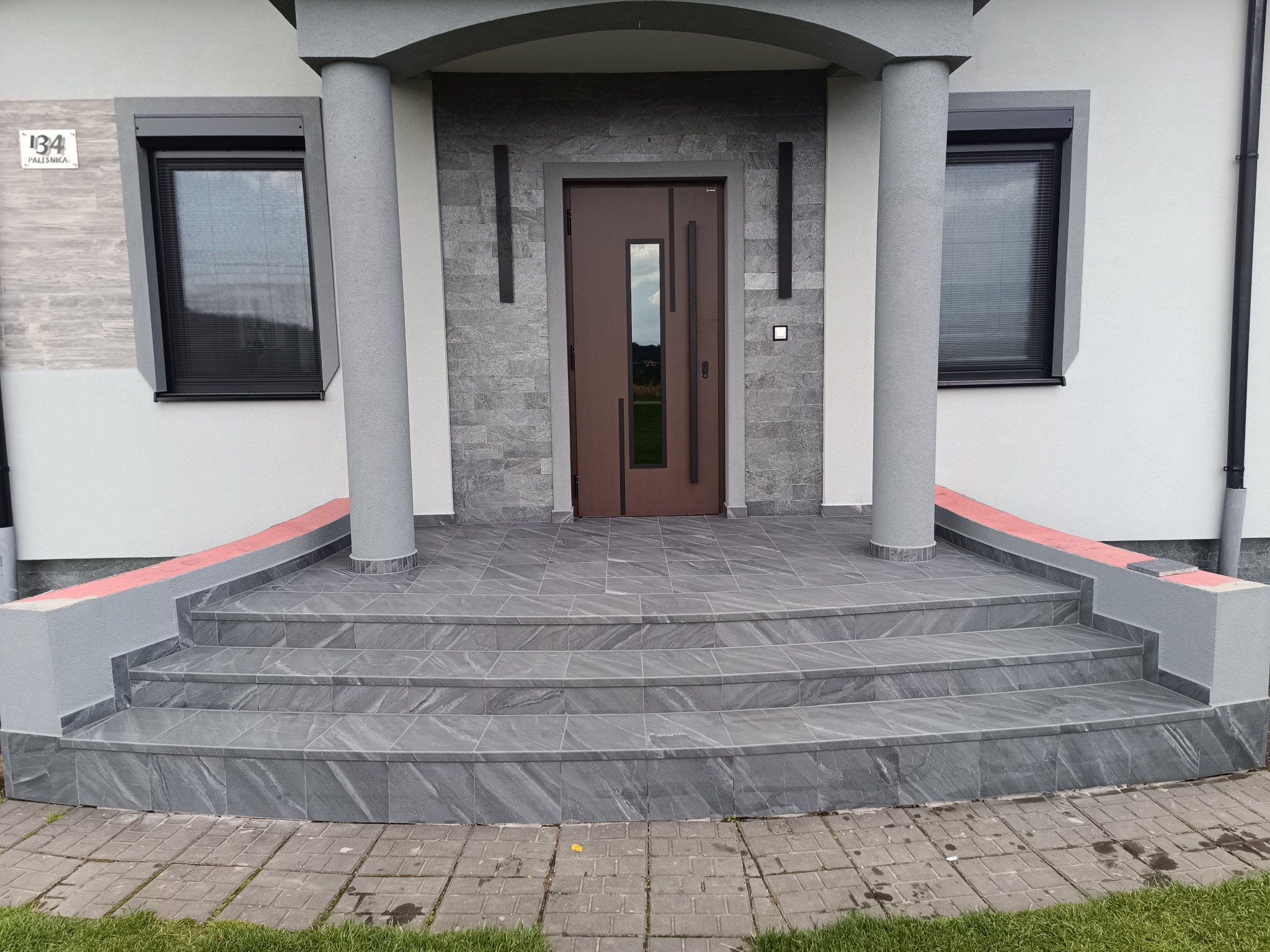 Wykończone schody wejściowe do domu z szarych płytek imitujących kamień, z ciemnobrązowymi drzwiami i szarą elewacją. Widoczne kolumny i numer domu.
