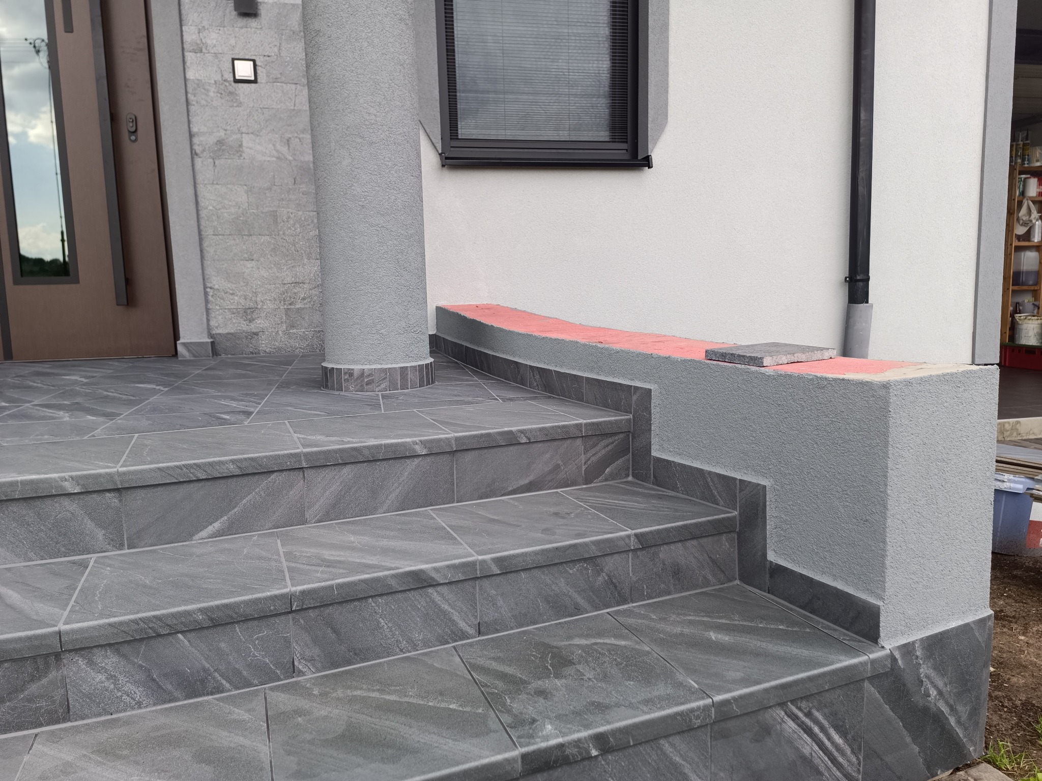 Zewnętrzne schody z szarych płytek imitujących kamień, prowadzące do wejścia. Widoczna kolumna i fragment elewacji z oknem.