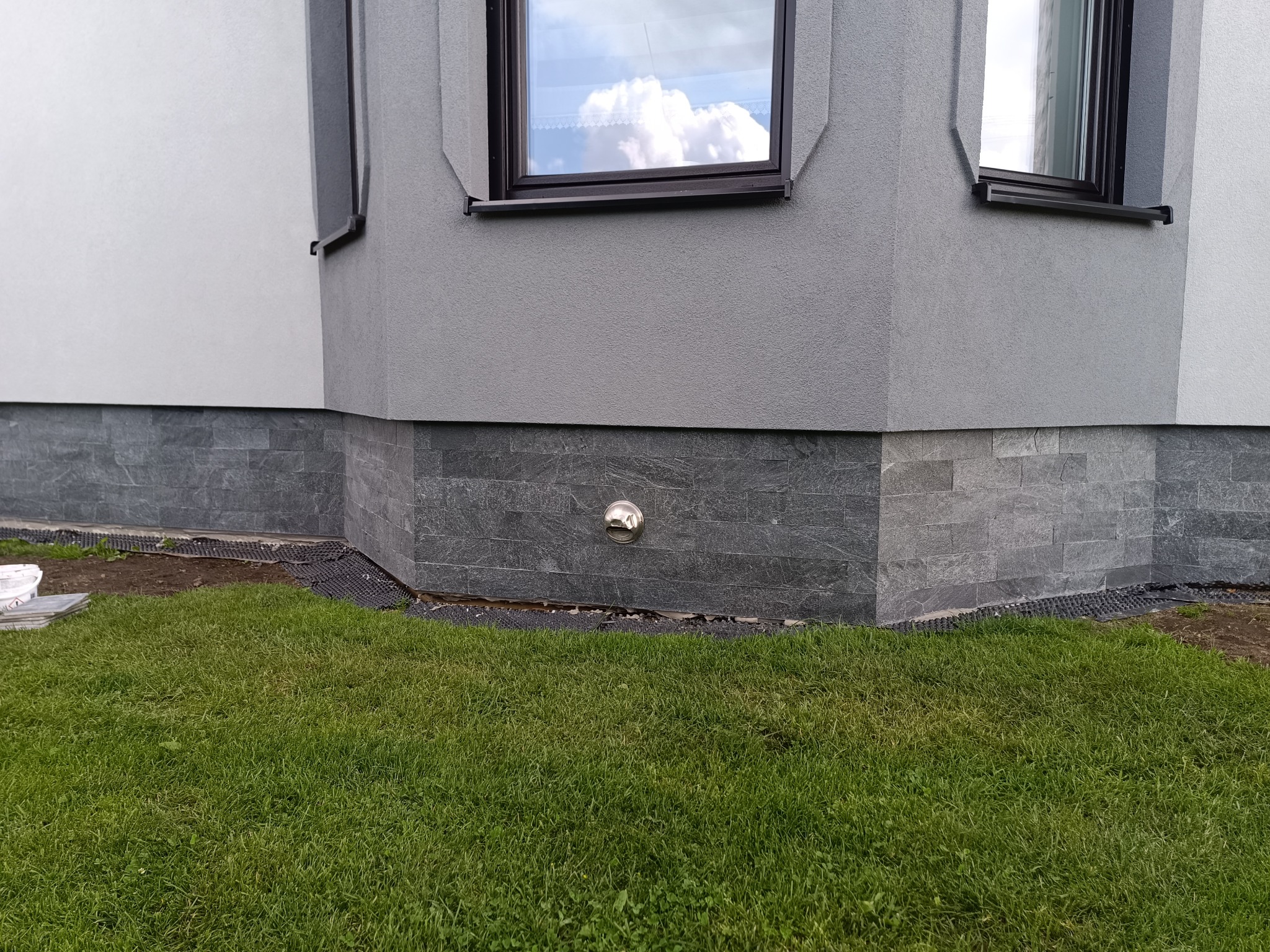 Wykończenie elewacji domu szarym kamieniem dekoracyjnym, widoczne okno z odbiciem chmur i trawnik. Detal srebrnej wentylacji. Nowoczesny design.