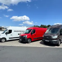 Flota trzech busów: biały z napisem 'Przeprowadzki', czerwony i brązowy, zaparkowane na parkingu w słoneczny dzień. Widoczny fragment nogi osoby.