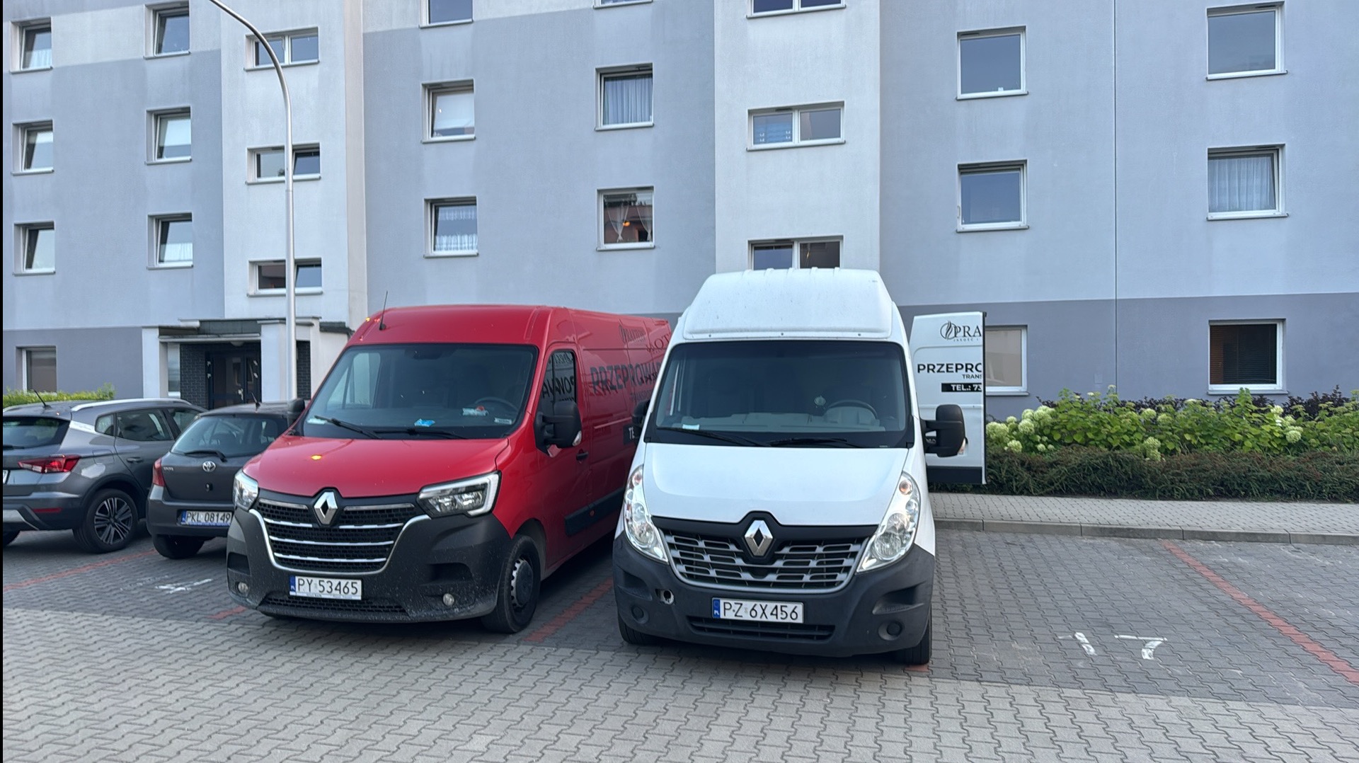 Dwa busy Renault Master (czerwony i biały) zaparkowane przed budynkiem mieszkalnym. Na czerwonym busie widoczne logo firmy przeprowadzkowej.