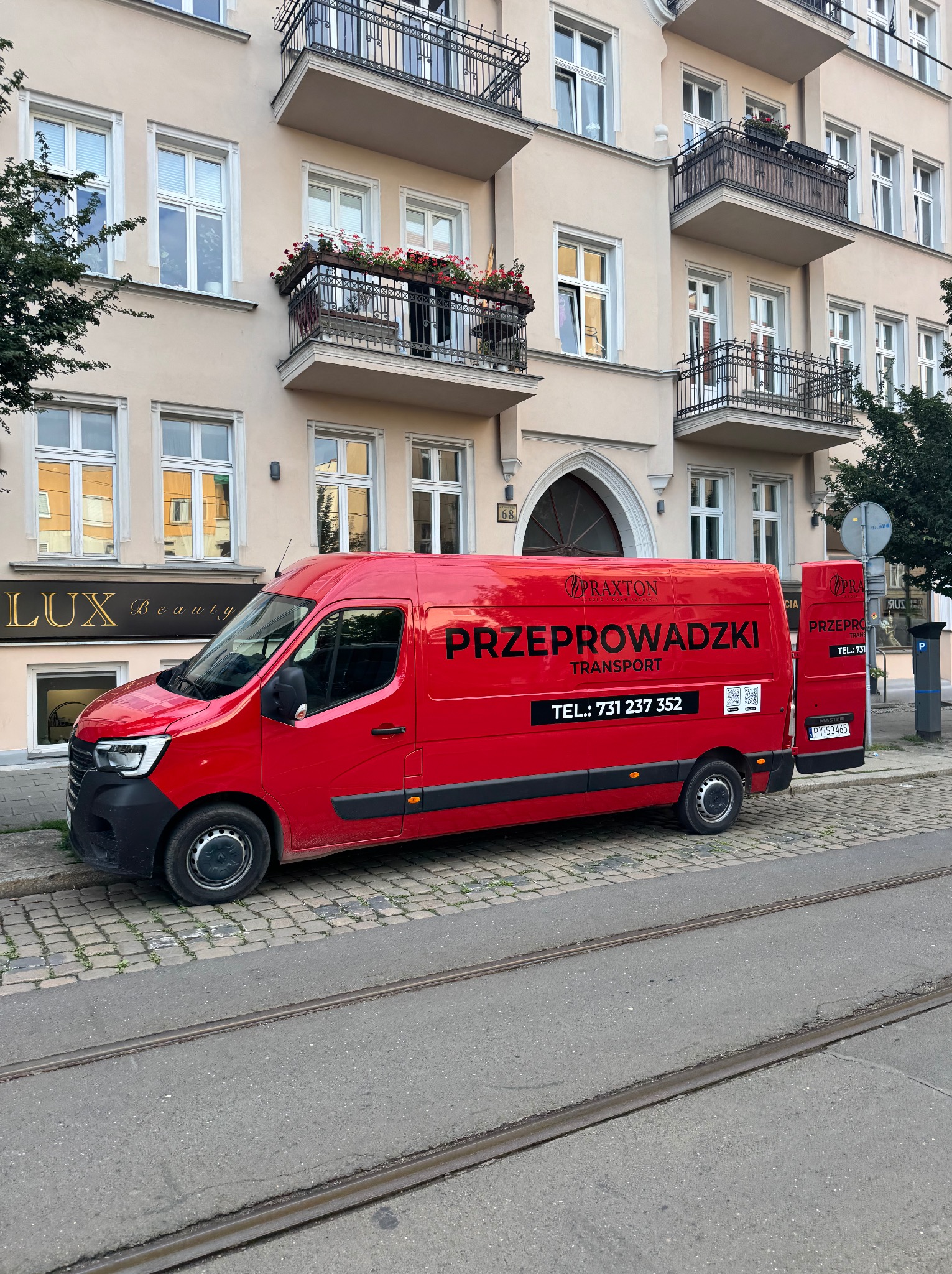 Czerwony bus z napisem 'Przeprowadzki' na tle kamienicy z balkonami, zaparkowany na ulicy z torami tramwajowymi. Otwarta tylna klapa. Widoczne logo firmy.