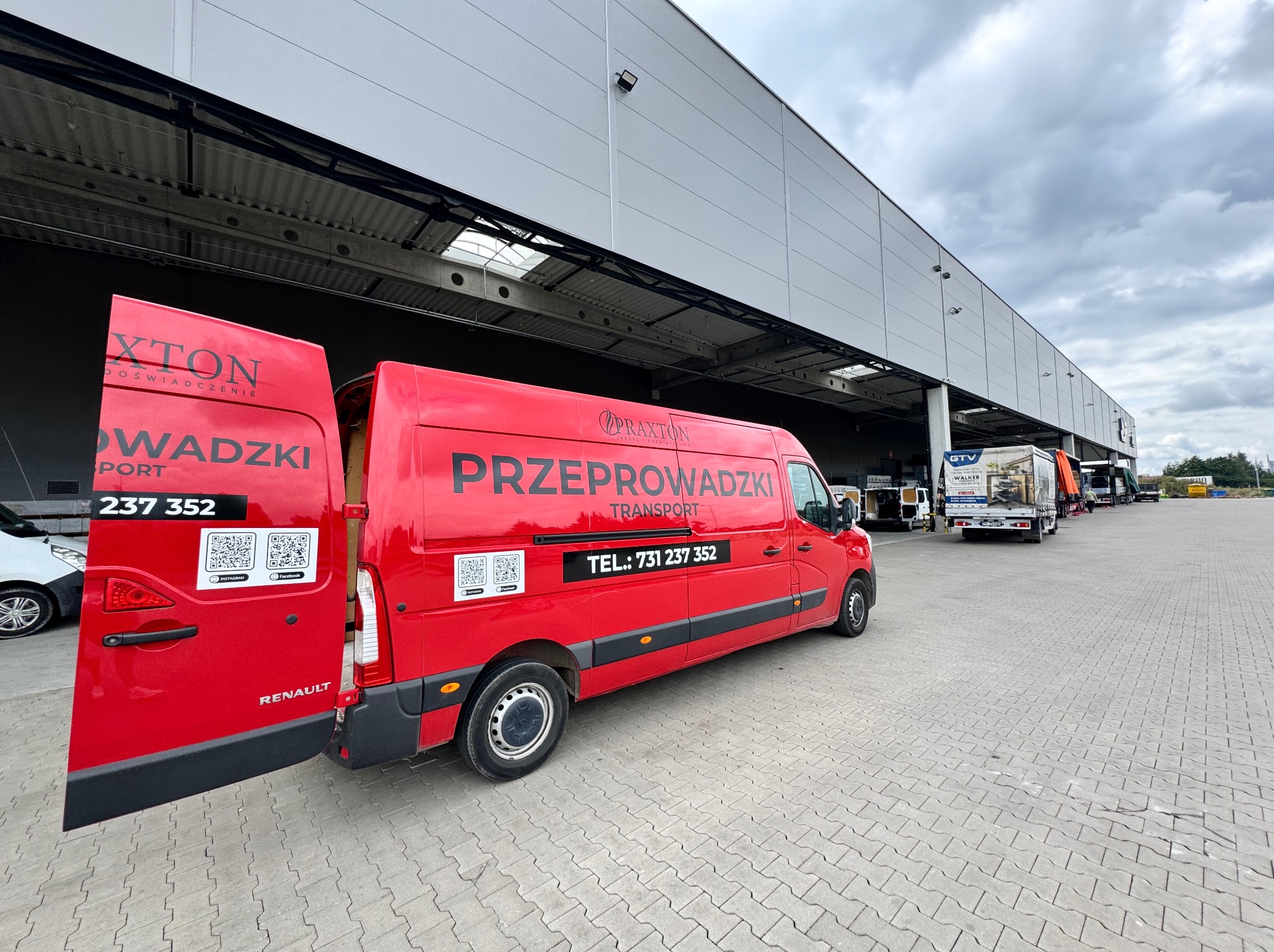 Czerwony bus Renault z otwartymi drzwiami, oklejony reklamą firmy przeprowadzkowej Praxton, zaparkowany na kostce brukowej przed magazynem. Widoczne numery telefonu i QR kody.