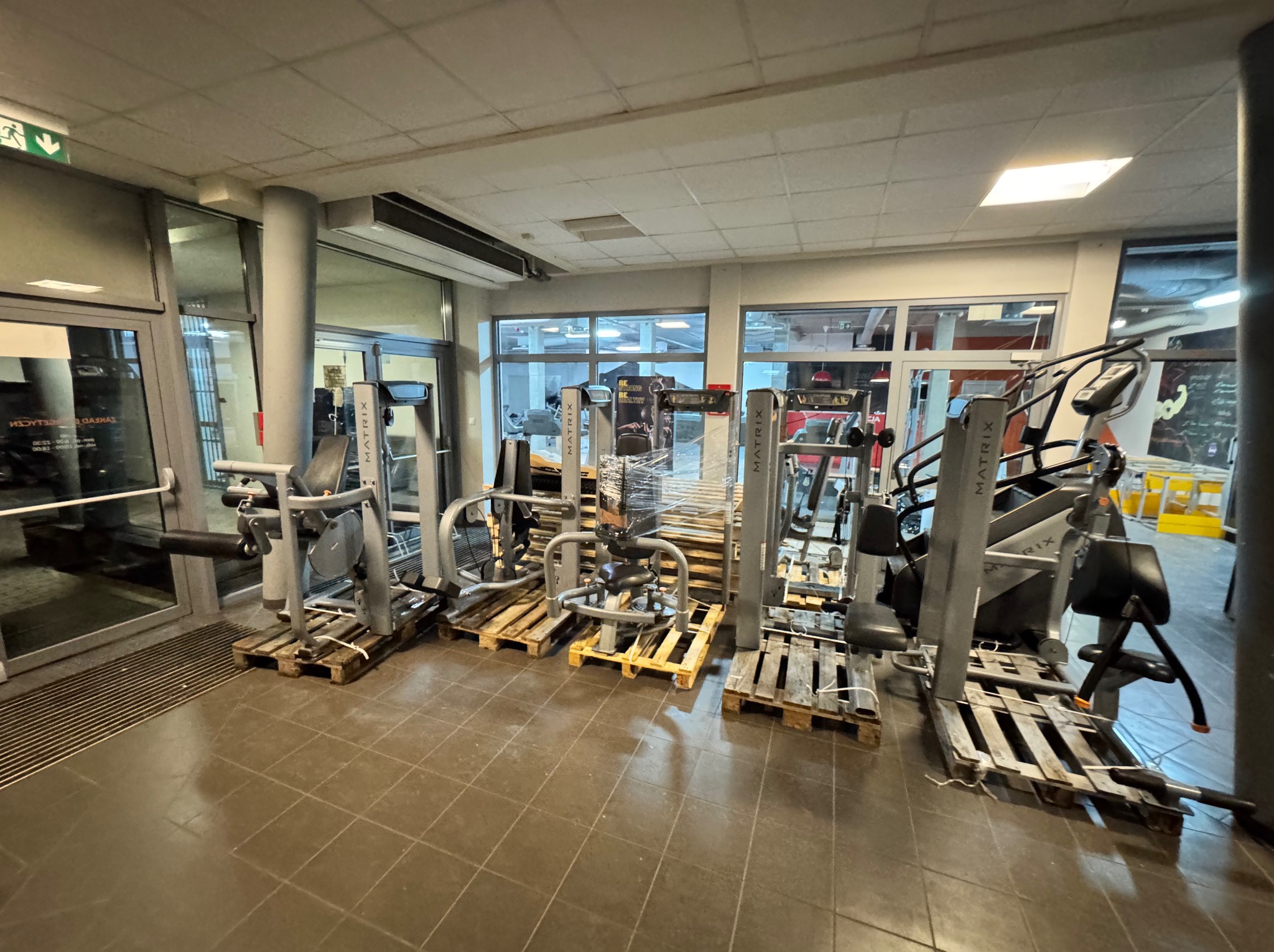 Transport siłowni: Sprzęt Matrix zabezpieczony na paletach, gotowy do przewozu. Widoczne bieżnie, maszyny siłowe, podłoga z płytek. Jasne wnętrze z oknami.