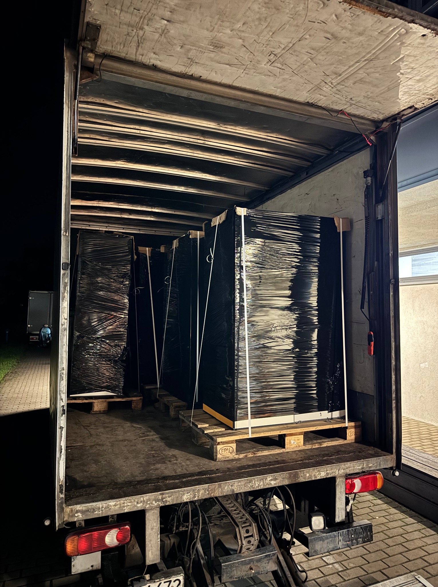 Wnętrze ciężarówki z paletami. Palety z ładunkiem zabezpieczone folią stretch. Widoczny fragment chodnika i ściana budynku. Transport nocą.