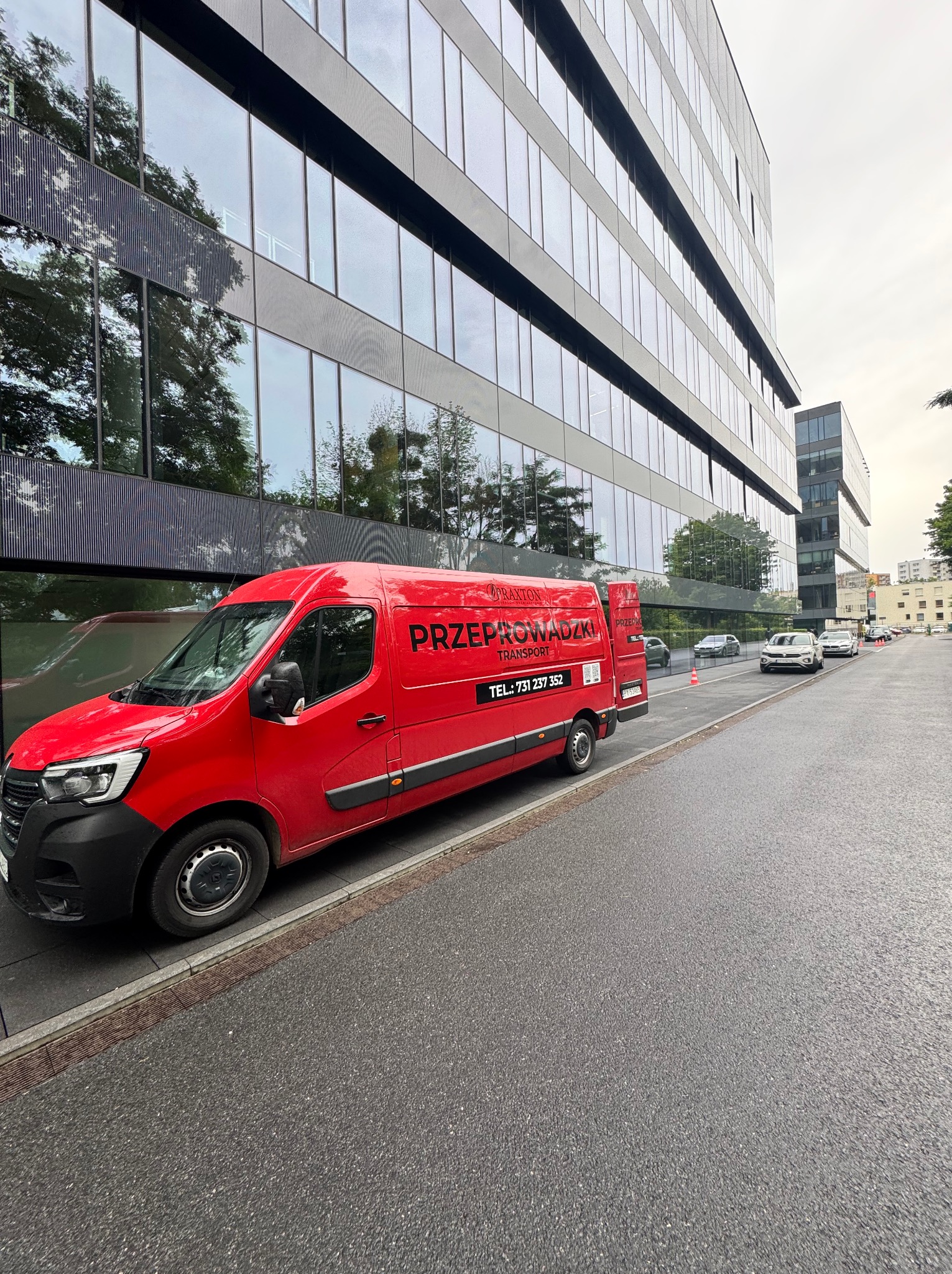 Czerwony bus z napisem 'Przeprowadzki Transport' zaparkowany przed nowoczesnym biurowcem z szklaną fasadą, odbijającą drzewa. Ulica z zaparkowanymi autami.