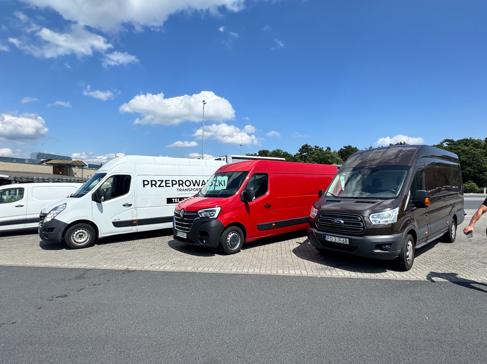 Flota trzech busów: biały z napisem 'Przeprowadzki', czerwony i brązowy, zaparkowane na parkingu w słoneczny dzień. Widoczny fragment nogi osoby.