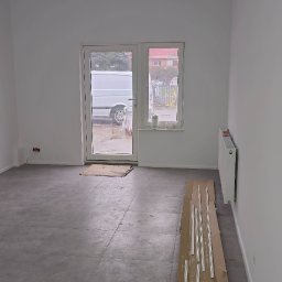 D&K Oleksandr Kruhliak - Pokój po remoncie: szare płytki podłogowe, białe ściany, grzejnik, listwy przypodłogowe na kartonie, widok na zewnątrz przez przeszklone drzwi z zaparkowanym białym vanem.