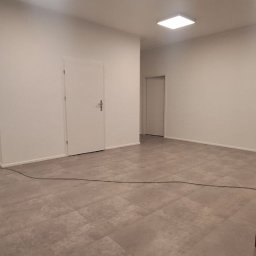 D&K Oleksandr Kruhliak - Wnętrze po odświeżeniu: świeżo pomalowane białe ściany, szara podłoga z płytek imitujących panele, widoczne drzwi wewnętrzne i stolik z krzesłami w rogu pomieszczenia.