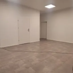 Wnętrze po odświeżeniu: świeżo pomalowane białe ściany, szara podłoga z płytek imitujących panele, widoczne drzwi wewnętrzne i stolik z krzesłami w rogu pomieszczenia.