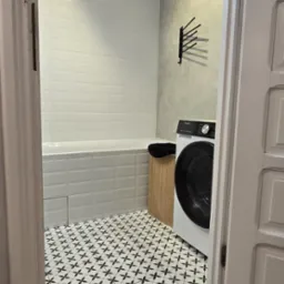 Wykończona łazienka z białą wanną, pralką i wzorzystą podłogą. Widoczne płytki ścienne, drewnopodobne panele podłogowe i oświetlenie sufitowe.