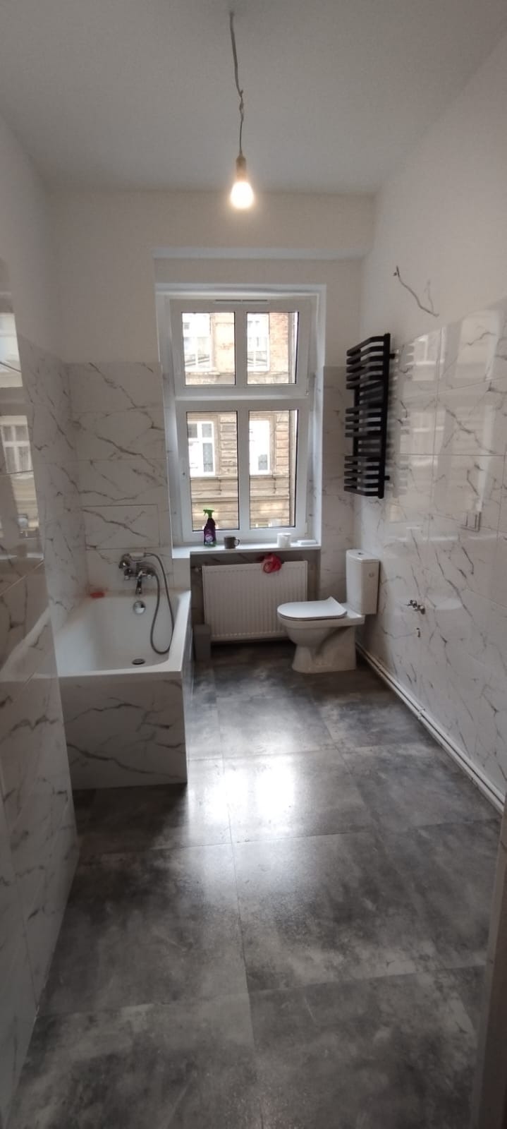 Wykończona łazienka z wanną obudowaną marmurową płytką, szarymi płytkami podłogowymi, grzejnikiem drabinkowym, toaletą i oknem z widokiem na budynek.