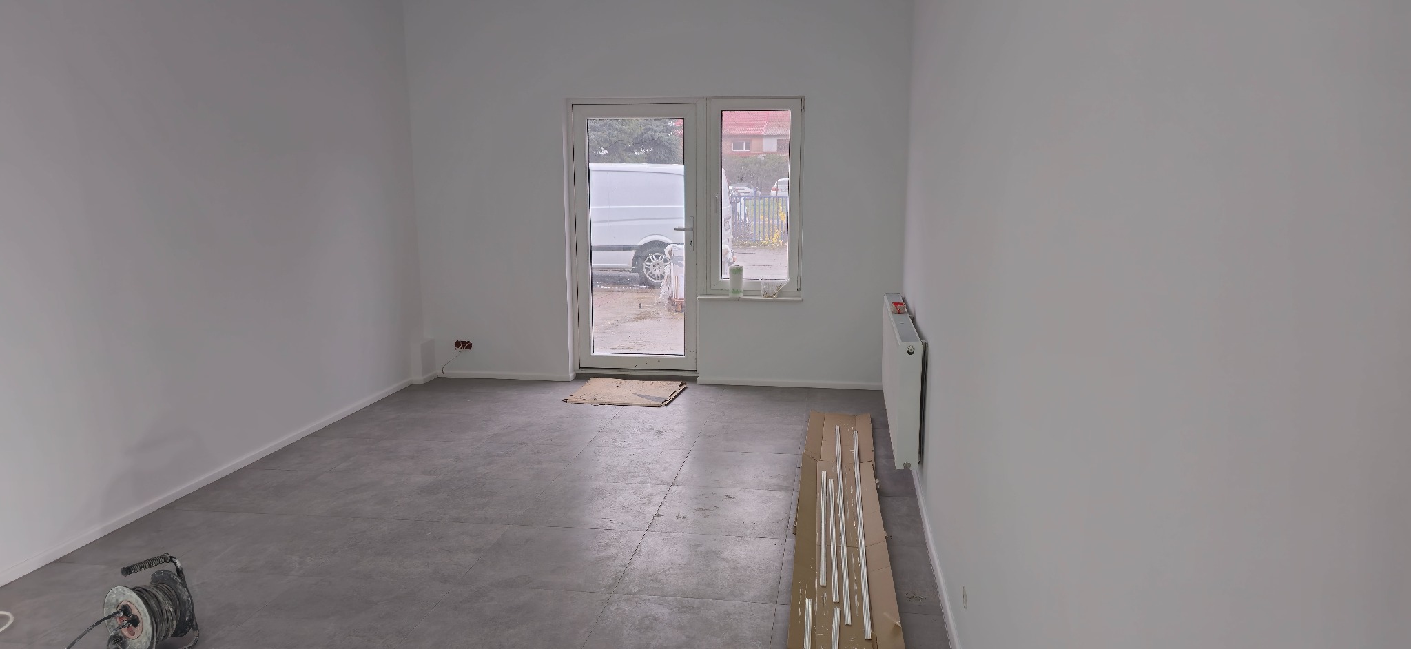 Pokój po remoncie: szare płytki podłogowe, białe ściany, grzejnik, listwy przypodłogowe na kartonie, widok na zewnątrz przez przeszklone drzwi z zaparkowanym białym vanem.