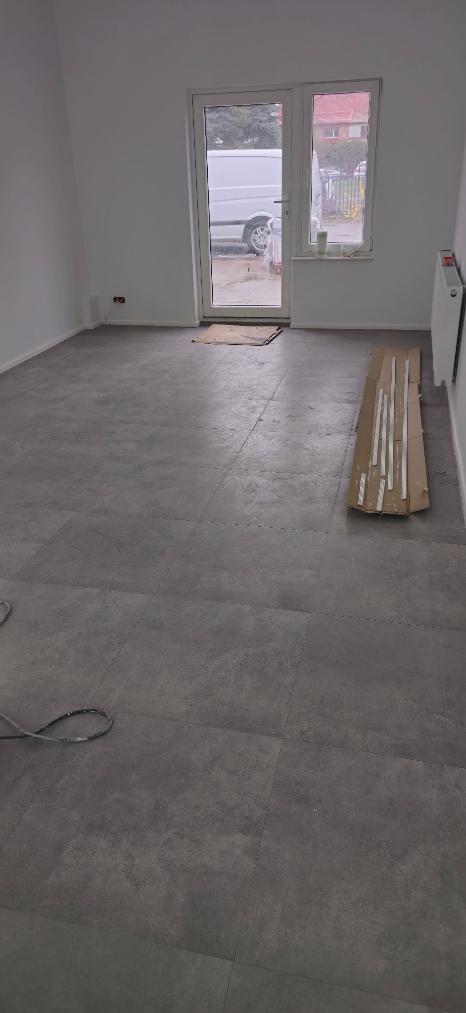 Szare panele podłogowe ułożone w nowym pomieszczeniu, widoczne listwy przypodłogowe w kartonie i kabel narzędzia na podłodze, białe ściany i drzwi balkonowe z widokiem na ulicę.