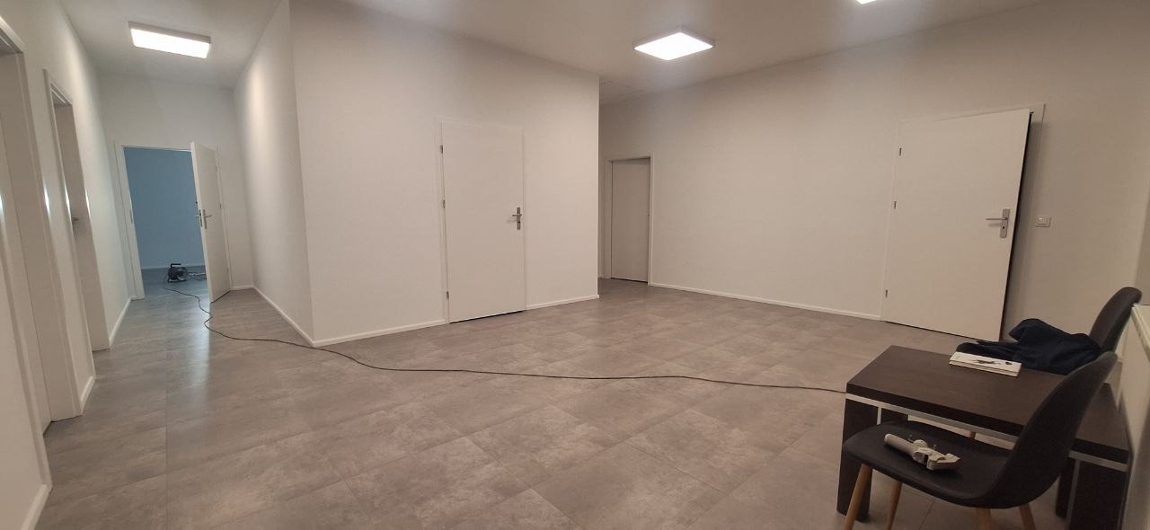 Wnętrze po odświeżeniu: świeżo pomalowane białe ściany, szara podłoga z płytek imitujących panele, widoczne drzwi wewnętrzne i stolik z krzesłami w rogu pomieszczenia.
