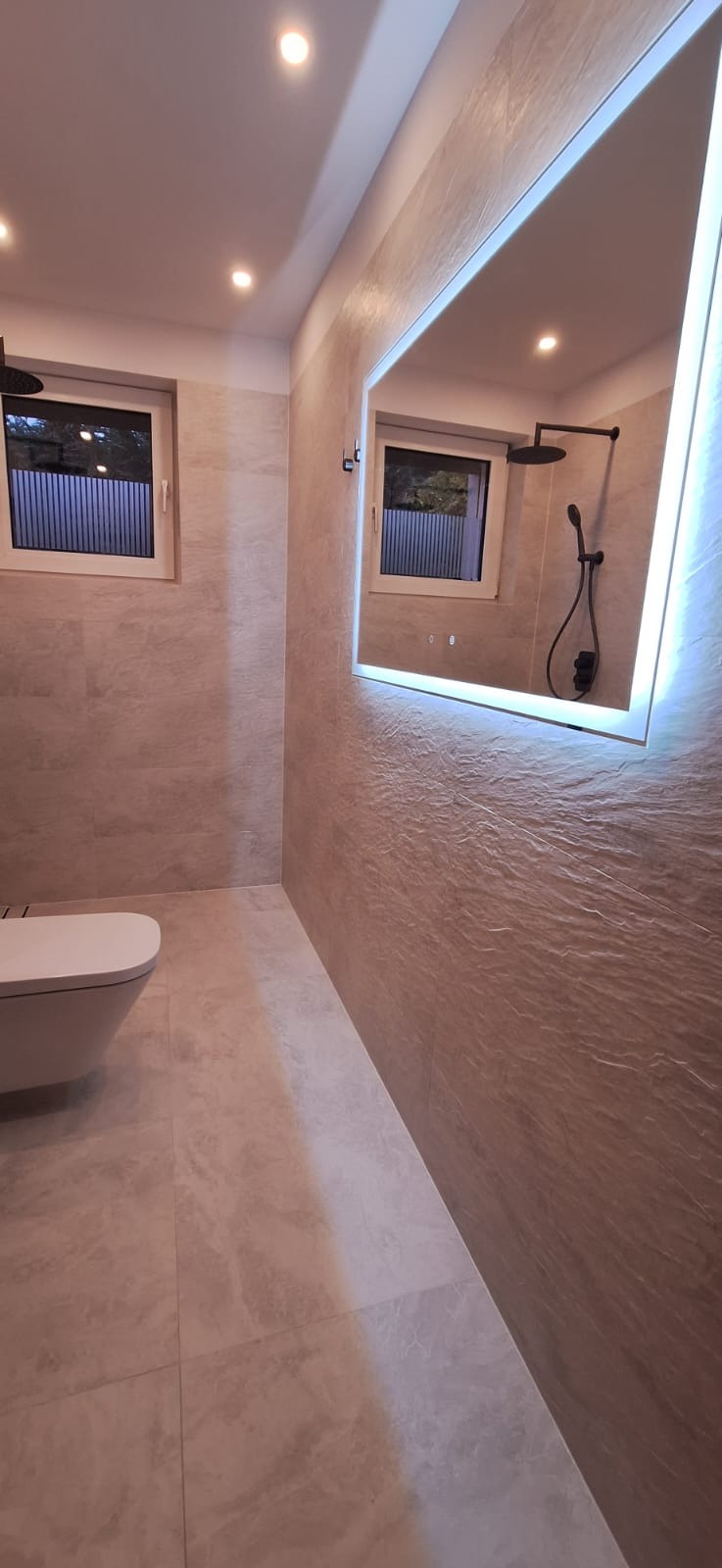 Nowoczesna łazienka z beżowymi płytkami imitującymi kamień, podświetlanym lustrem i wiszącą miską WC, widok na okno i czarną deszczownicę odbijającą się w lustrze.