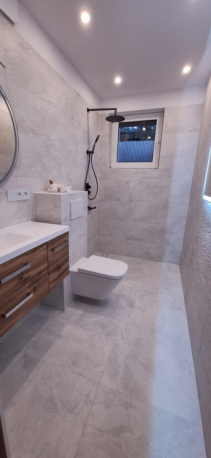 Nowoczesna łazienka z szarymi płytkami imitującymi marmur, drewnianą szafką pod umywalką, wiszącą toaletą i czarną armaturą prysznicową.