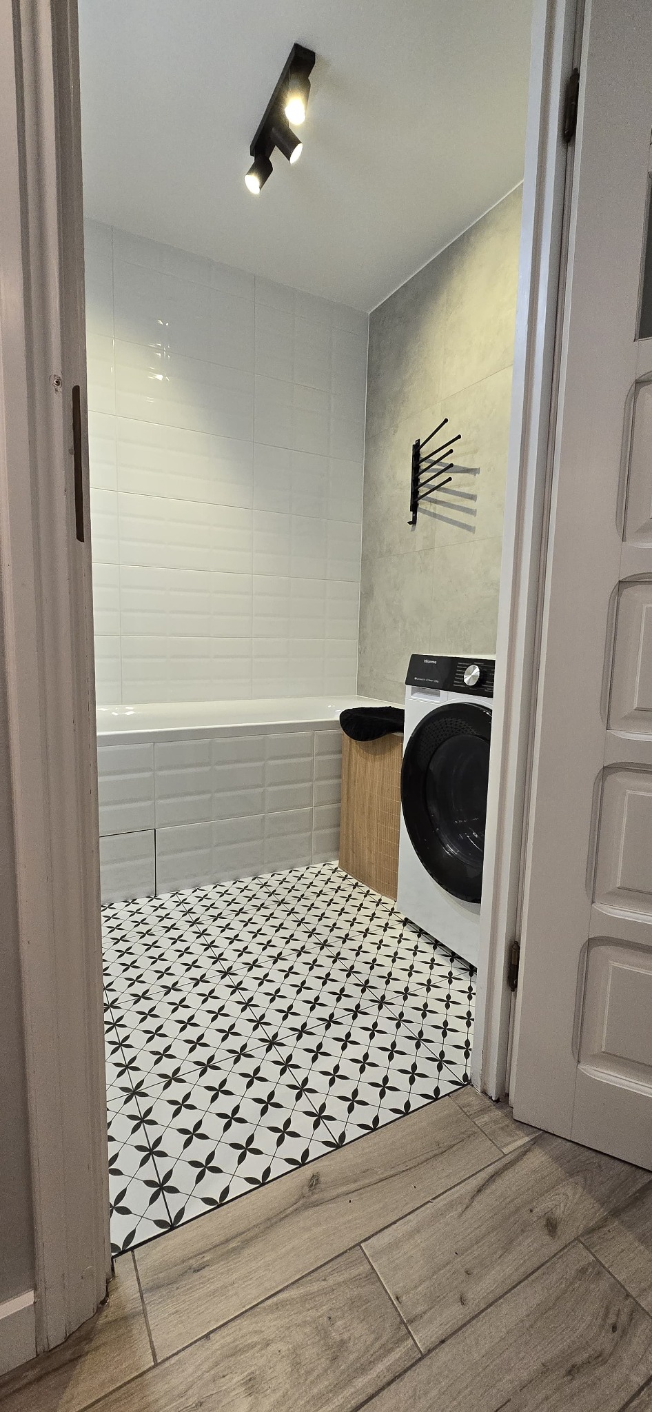 Wykończona łazienka z białą wanną, pralką i wzorzystą podłogą. Widoczne płytki ścienne, drewnopodobne panele podłogowe i oświetlenie sufitowe.