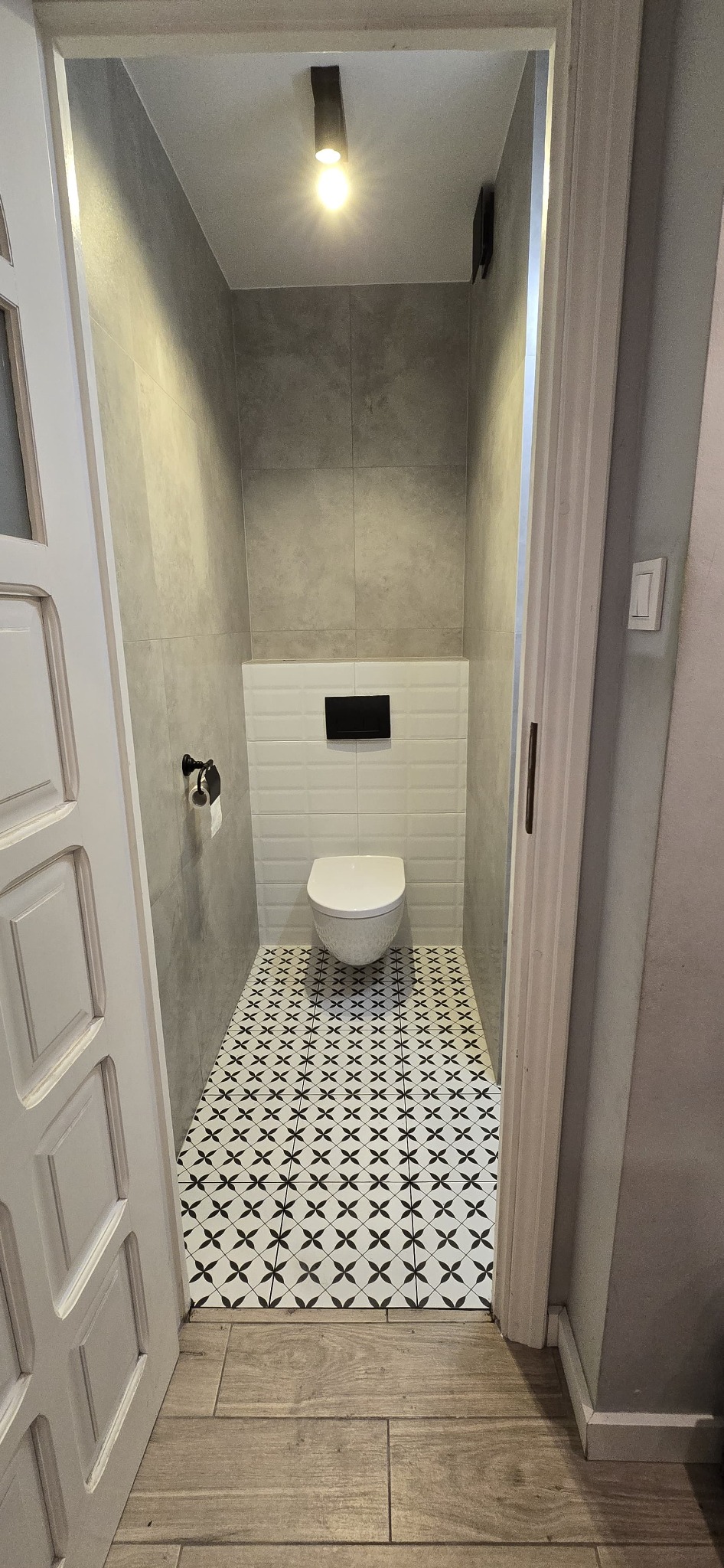 Mała toaleta w szarościach z białą ceramiką i wzorzystą podłogą. Widok zza uchylonych, białych drzwi.