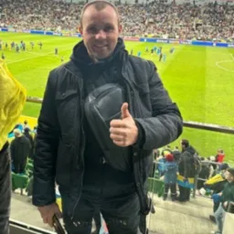 Mężczyzna w czarnej kurtce i saszetce nerkowej pozuje na tle trybun stadionu, pokazując kciuk w górę.