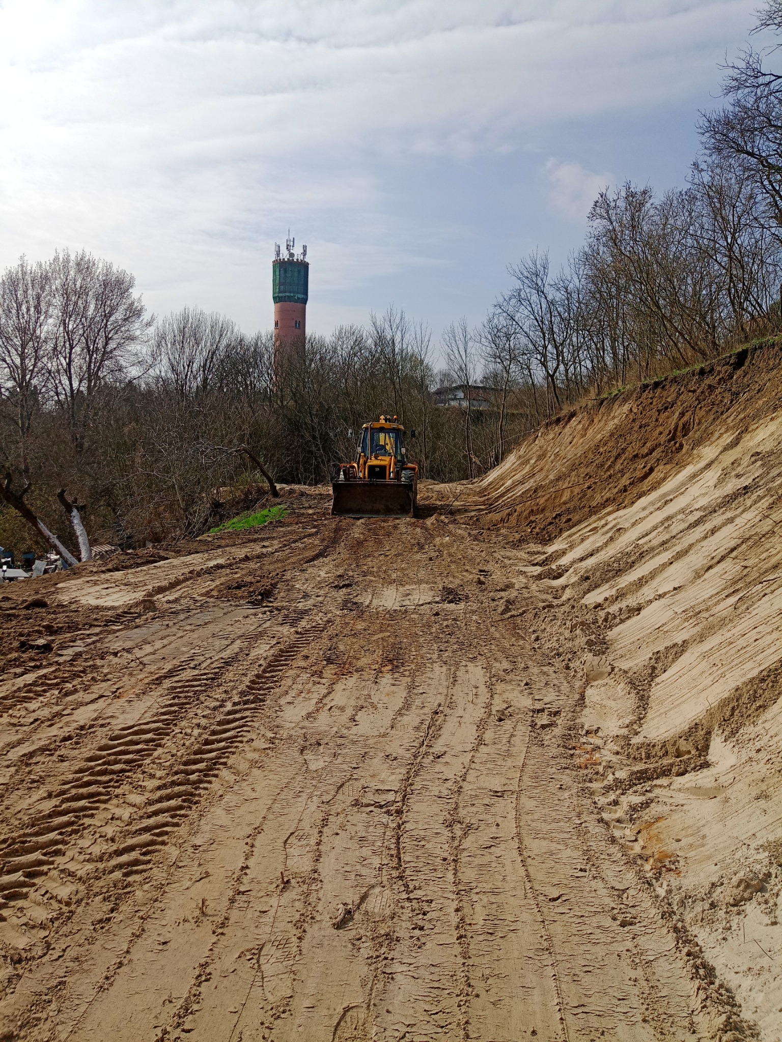 Żółta koparko-ładowarka na piaszczystym terenie z widocznymi śladami opon, w tle drzewa i wieża ciśnień z antenami.