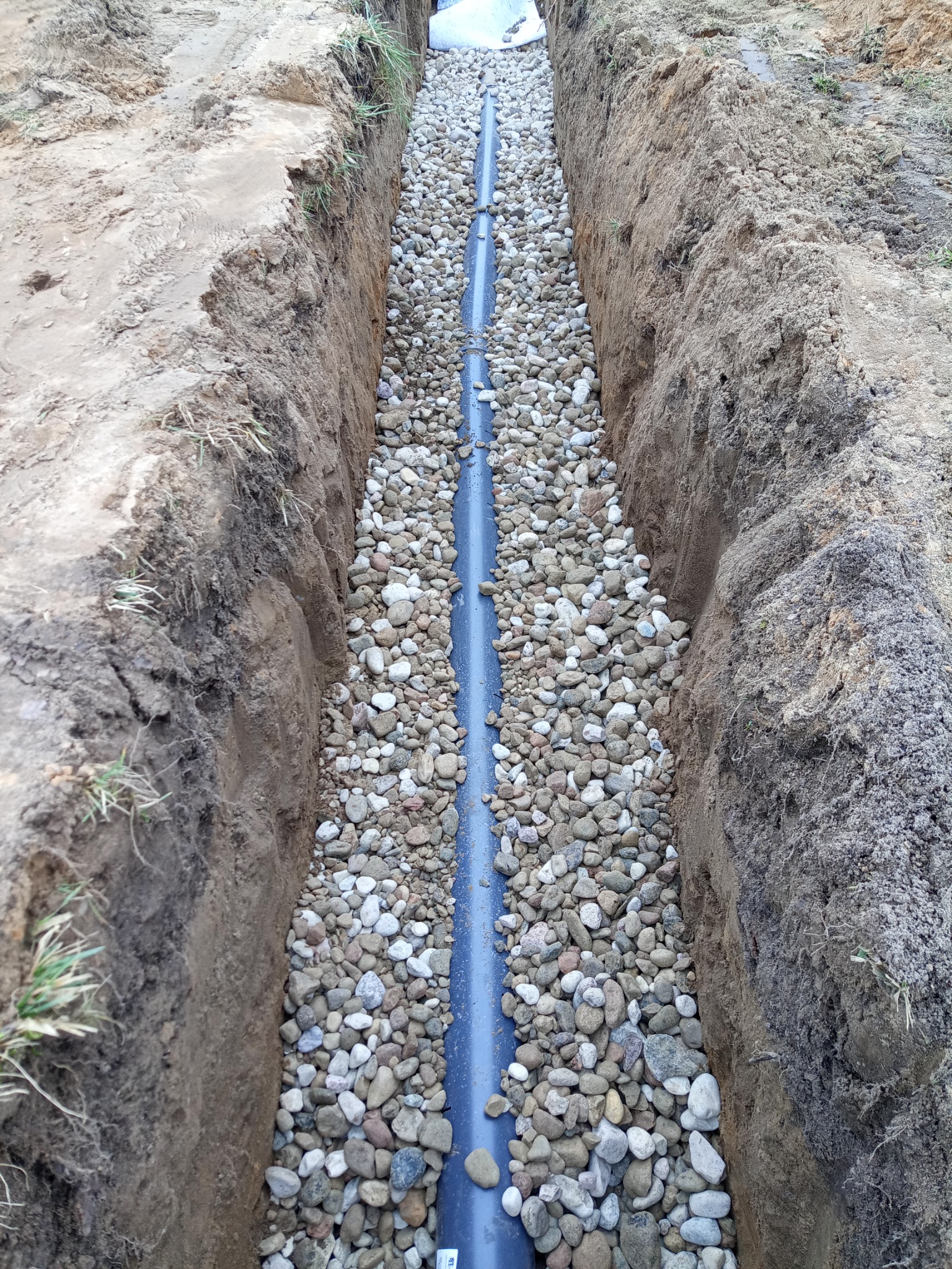 Wąski wykop w ziemi z ułożoną rurą i obsypany drobnymi kamieniami, widoczne ślady niedawnych prac ziemnych.