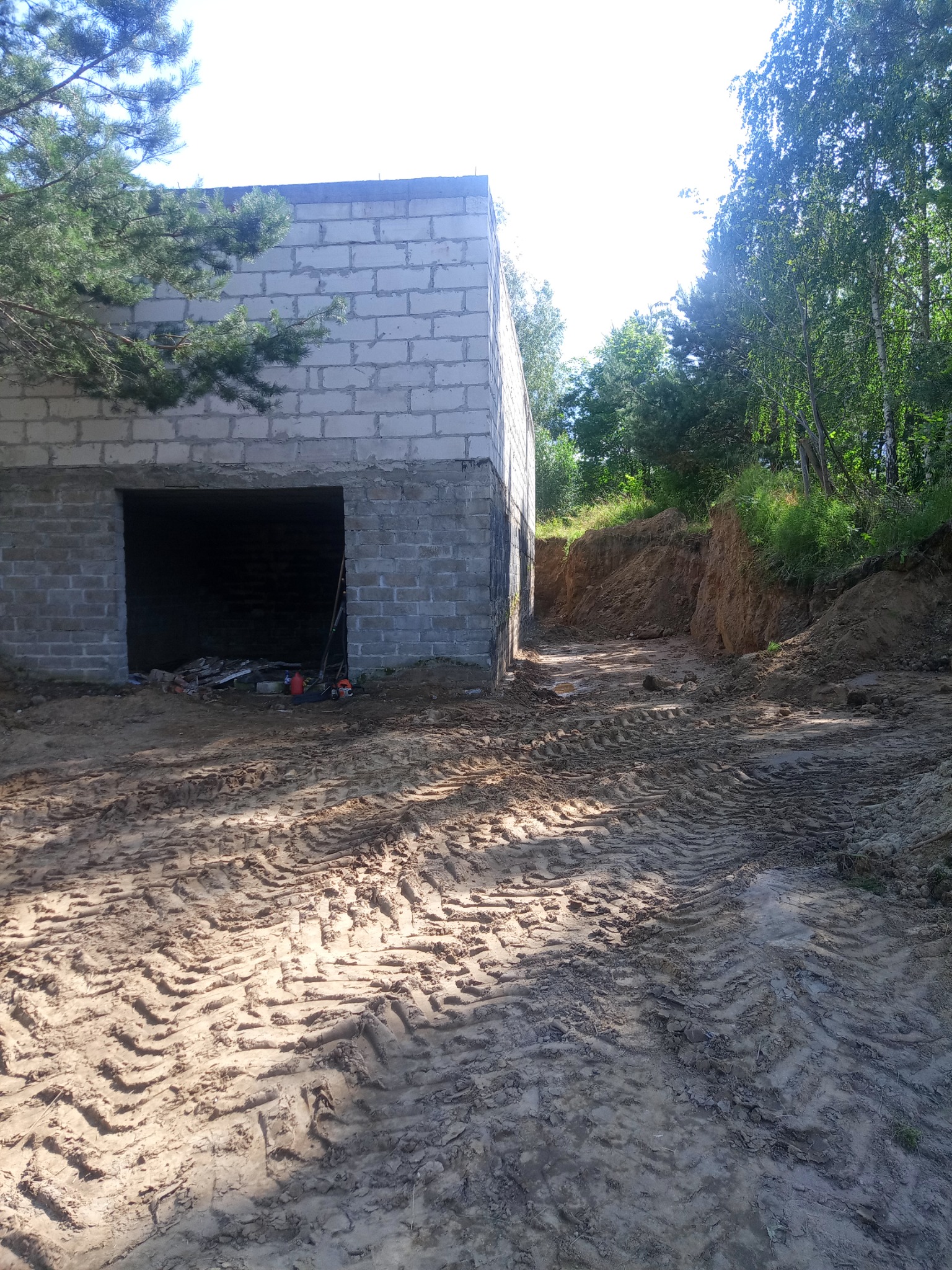 Surowy budynek z pustaków na nierównym, piaszczystym terenie z wyraźnymi śladami opon, częściowo otoczony drzewami i wykopem ziemnym.