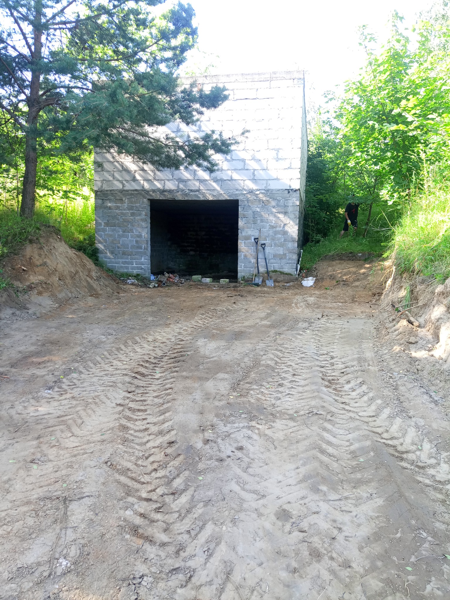 Surowy budynek z szarych bloczków z otwartym wjazdem, widoczne ślady opon na ziemi, dwie łopaty opierają się o ścianę, teren częściowo wyrównany, w tle zieleń drzew.