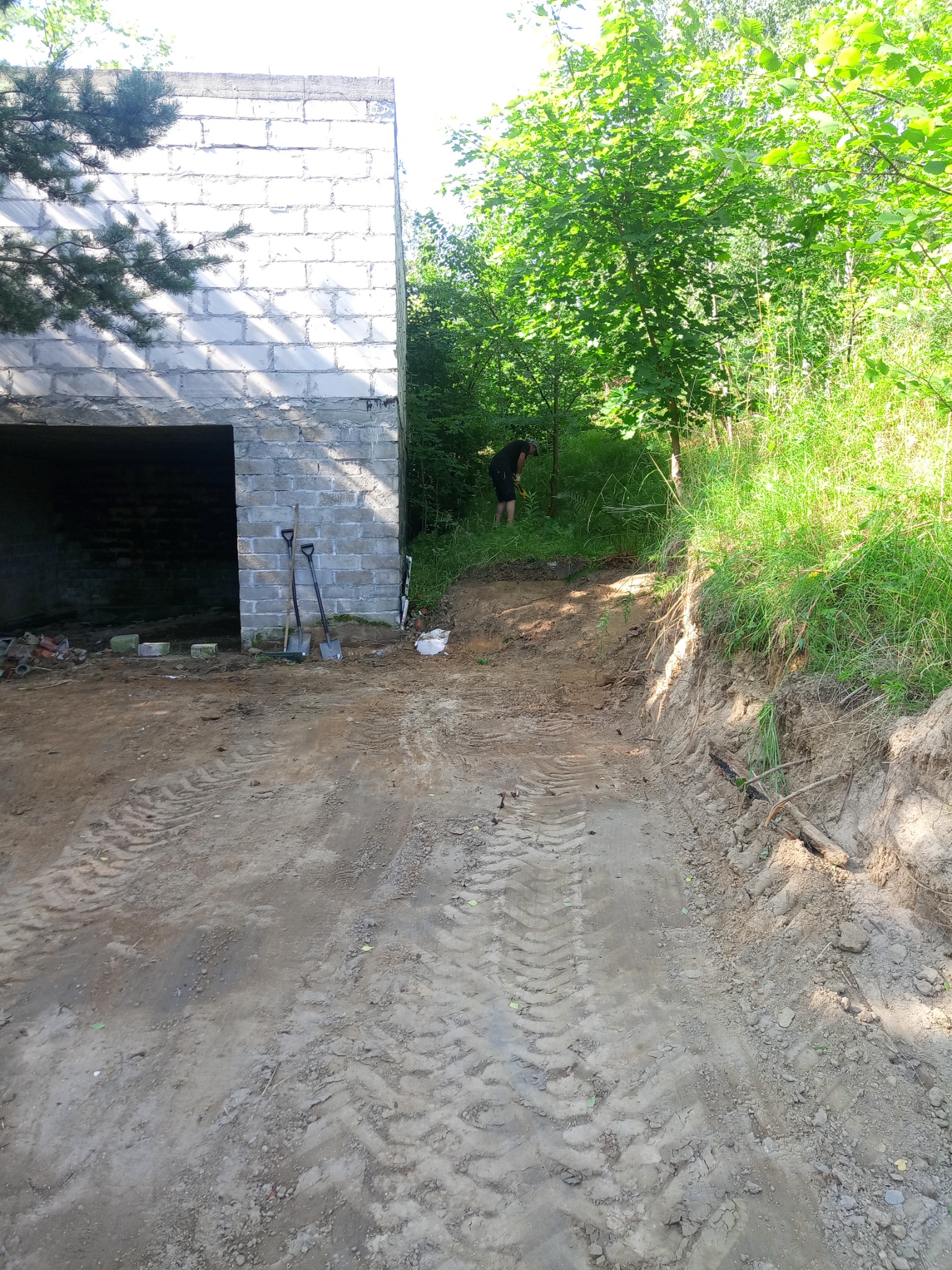 Przygotowanie terenu pod budowę: widok na fundamenty w stanie surowym, dwie łopaty opierają się o ścianę, w tle osoba wyrównuje teren w zaroślach, ślady opon na ubitej ziemi.