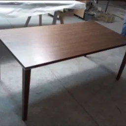 Stół drewniany o prostej, minimalistycznej konstrukcji z ciemnego drewna, sfotografowany w warsztacie stolarskim, z widocznymi elementami wyposażenia w tle.