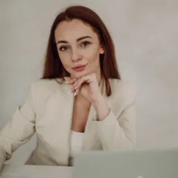 Portret kobiety w beżowym żakiecie, opierającej brodę na dłoni, z laptopem i dokumentami na biurku.