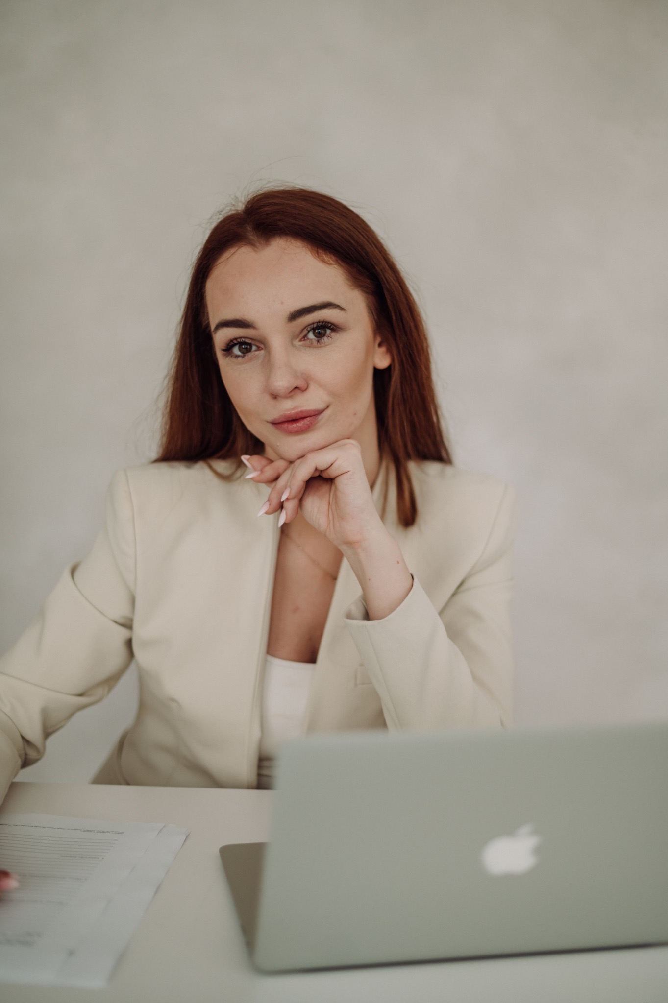 Portret kobiety w beżowym żakiecie, opierającej brodę na dłoni, z laptopem i dokumentami na biurku.